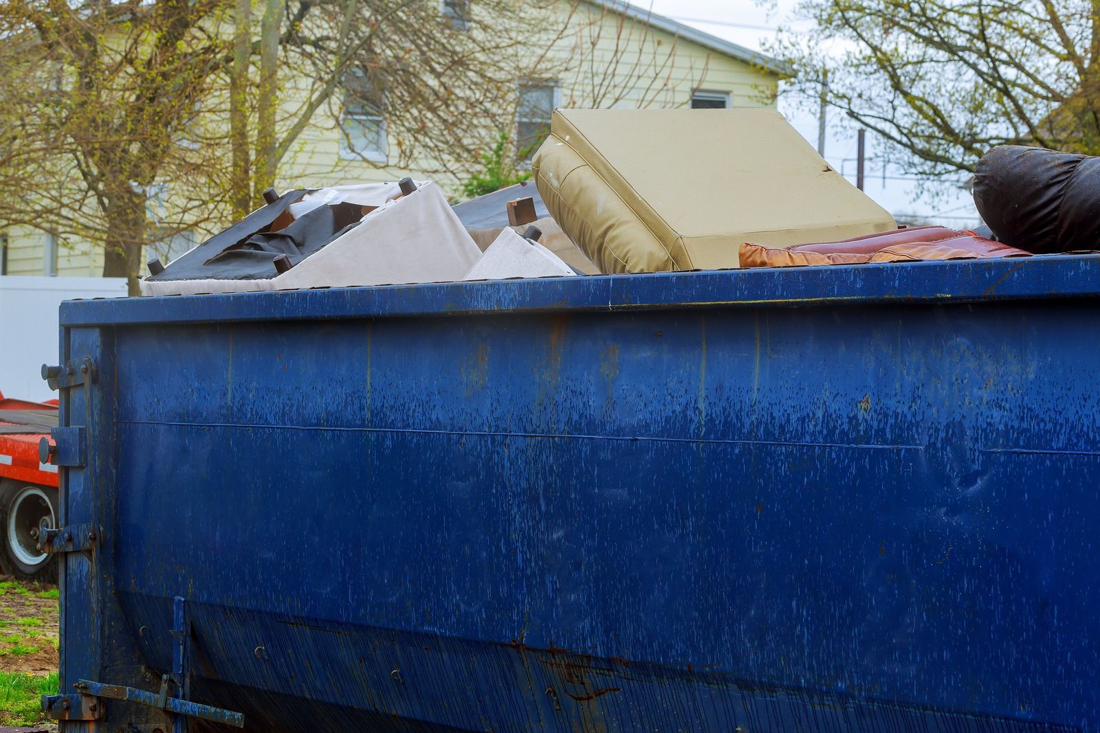 Blue dumpster filled with debris and a rolled carpet outside a house