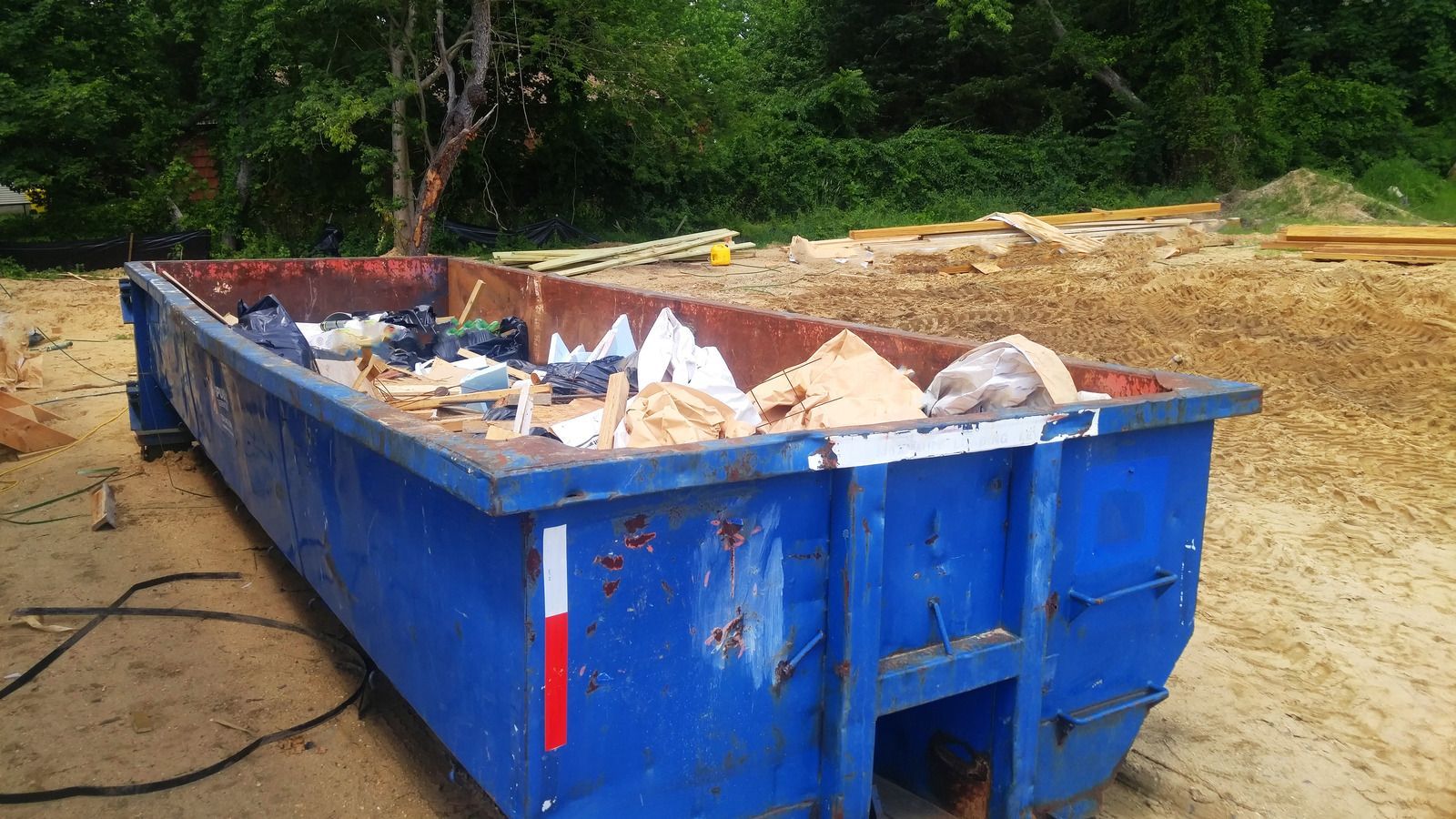 Blue dumpster filled with construction debris on a dirt lot near trees and a sandy work area