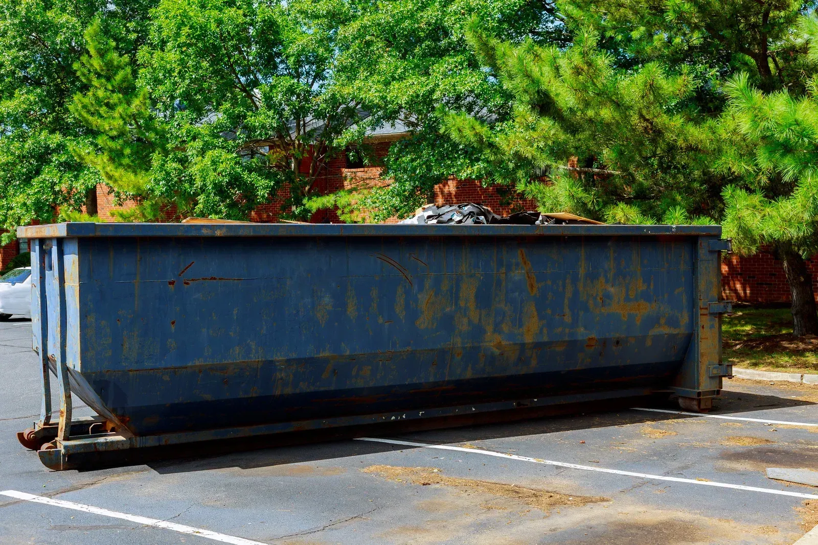 Large blue dumpster in a parking lot beside green trees, with rust and wear visible.