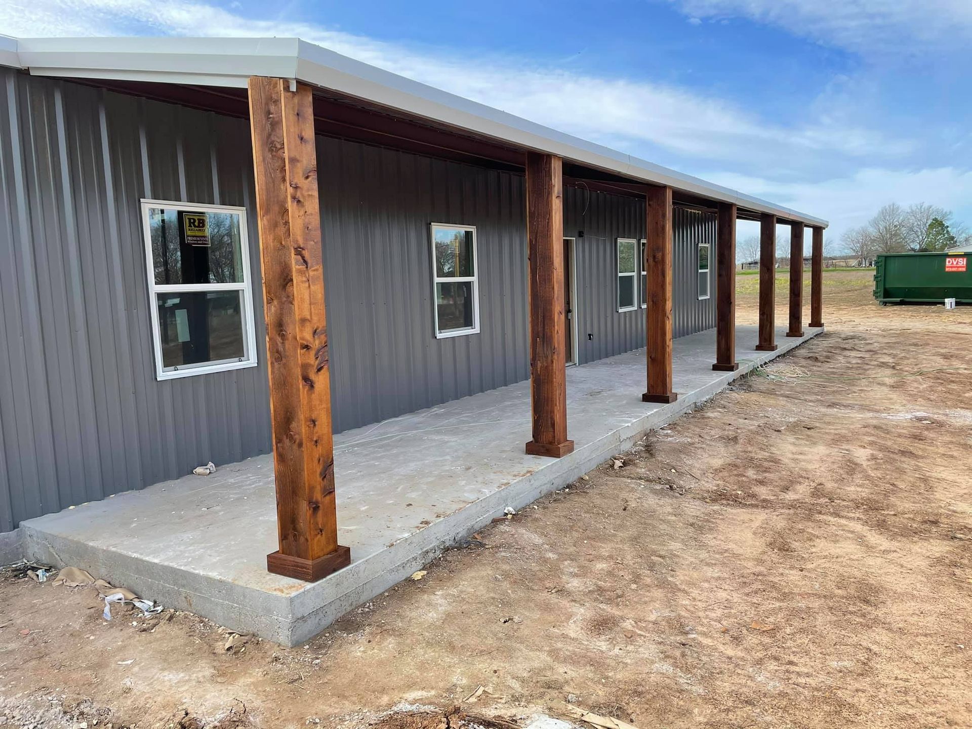 New gray metal building with wooden porch posts and concrete walkway on a dirt lot
