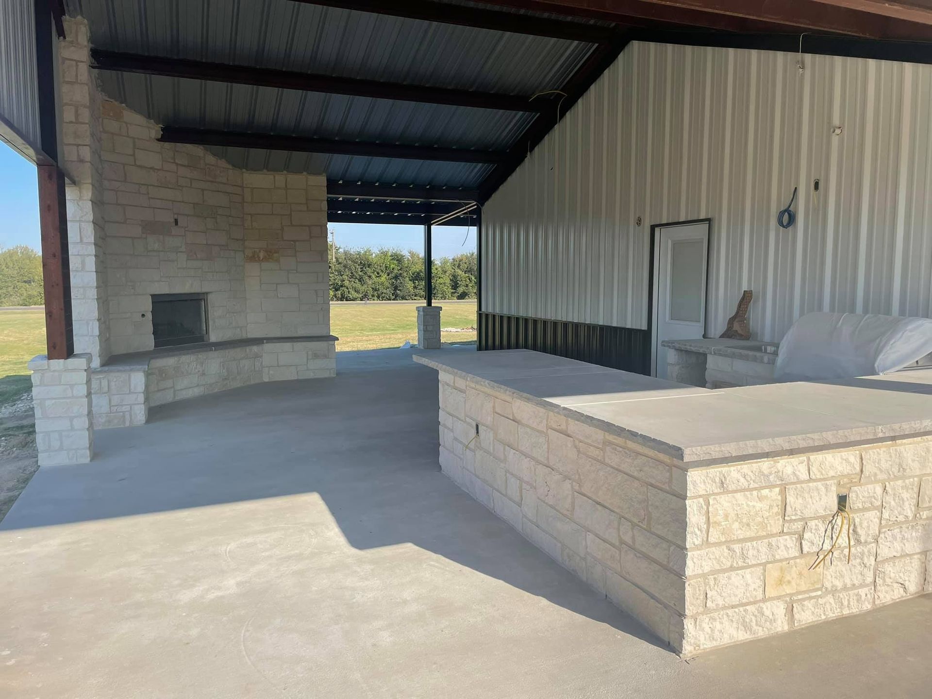 Covered outdoor patio with stone benches, concrete floor, and a white metal wall overlooking a grassy field