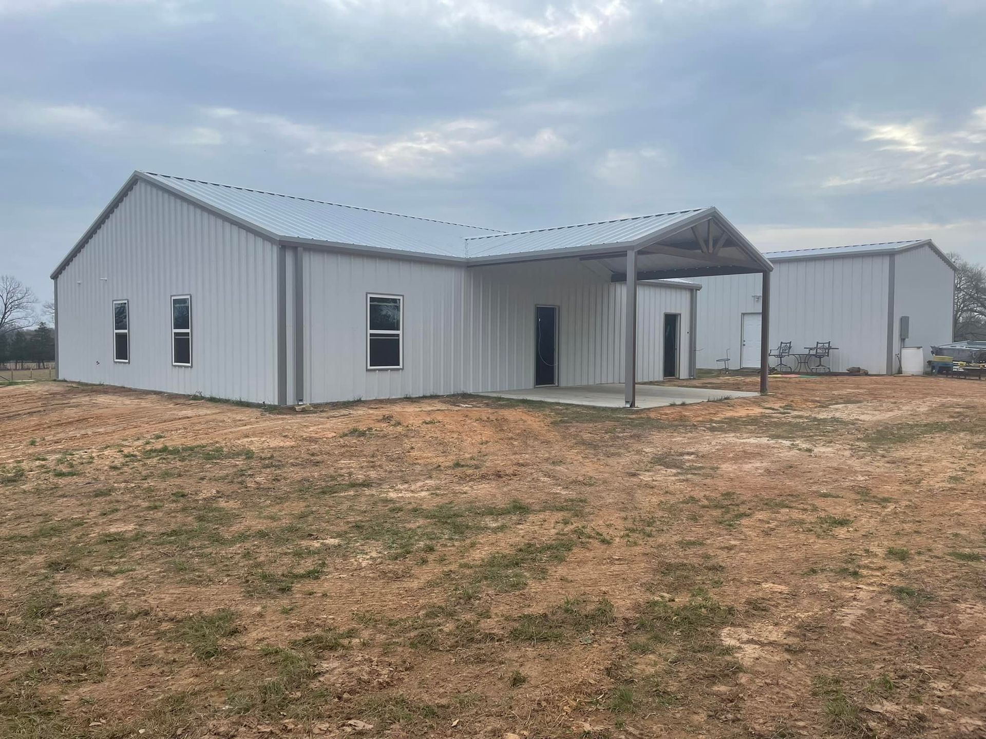 Small gray metal house with a covered porch on a bare dirt lot under a cloudy sky.