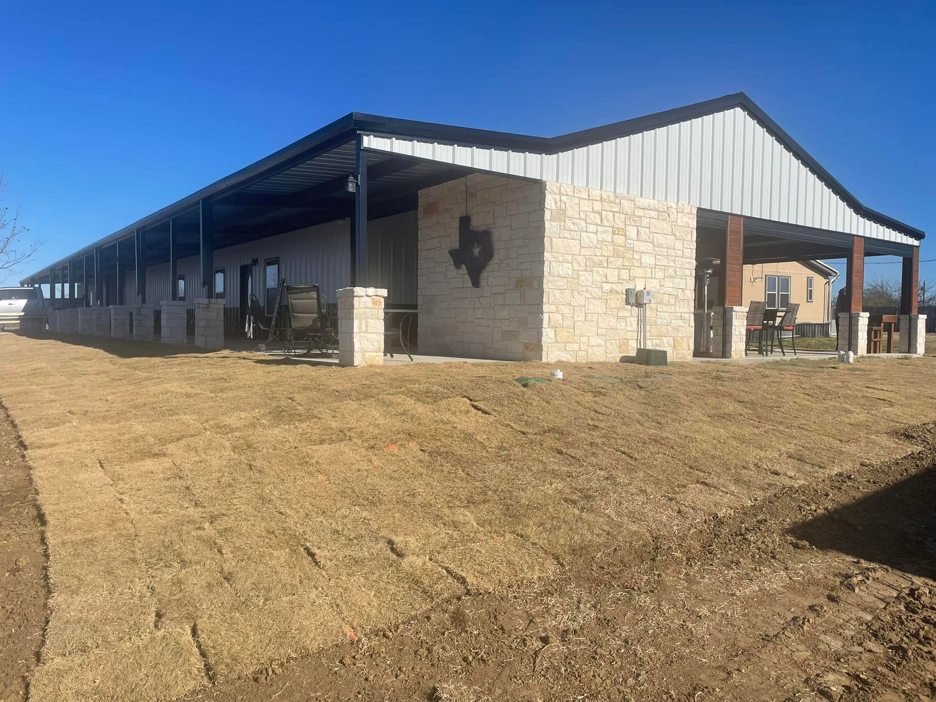 Long stone-and-metal ranch building with a covered porch on a dry grassy lot under a blue sky