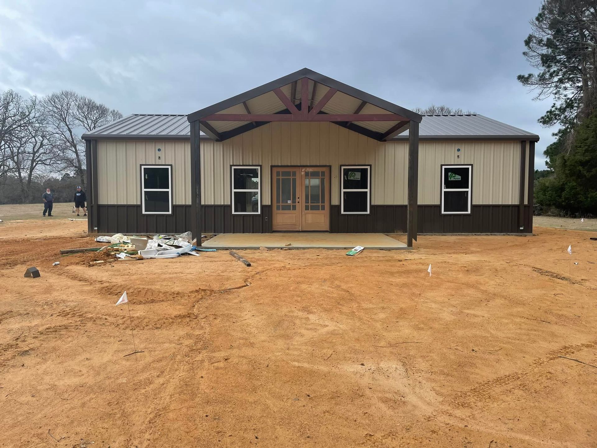 New beige house with dark trim and a front porch on a bare dirt lot under an overcast sky