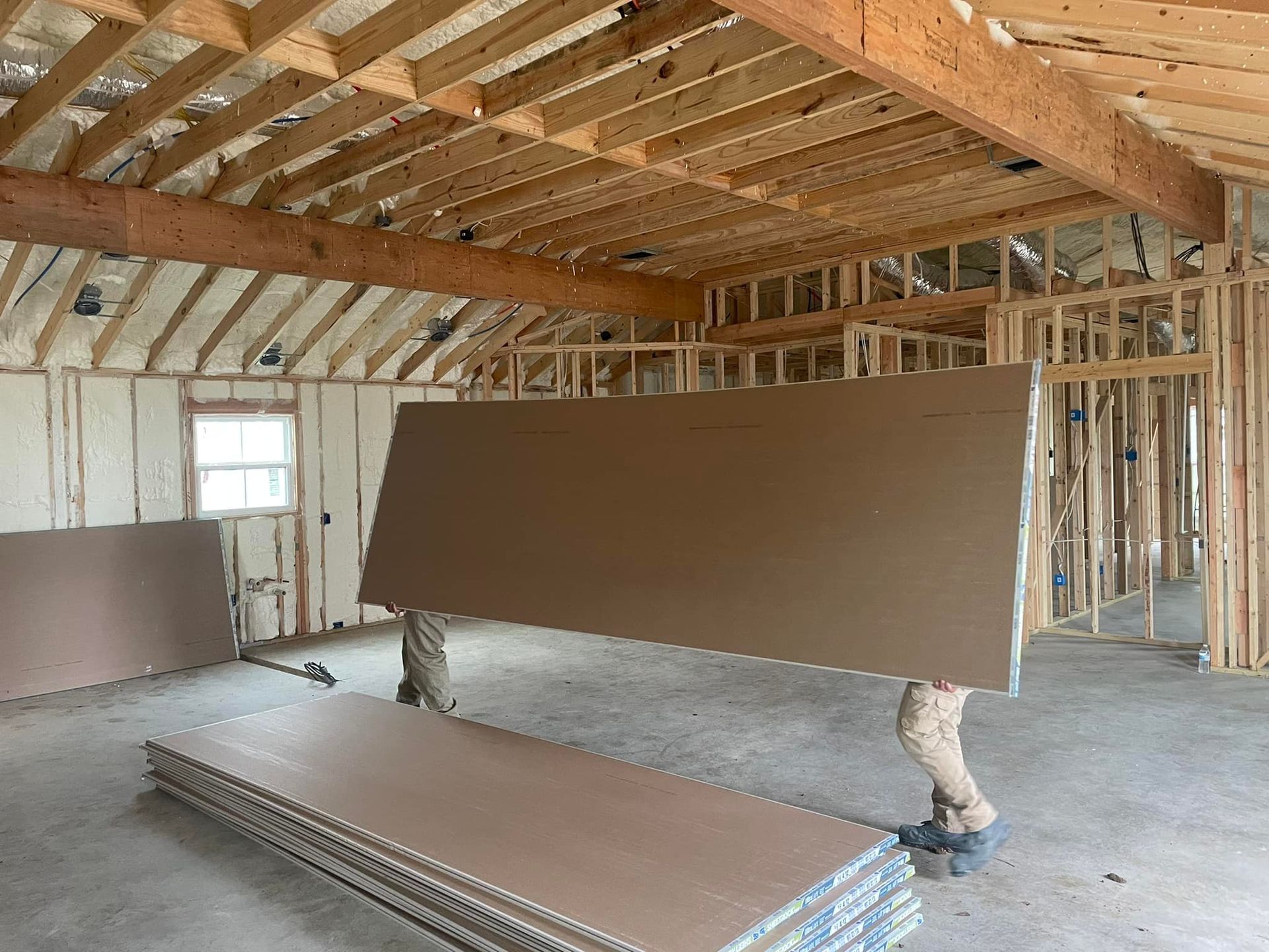 Construction site with workers lifting a large drywall sheet inside a framed house.