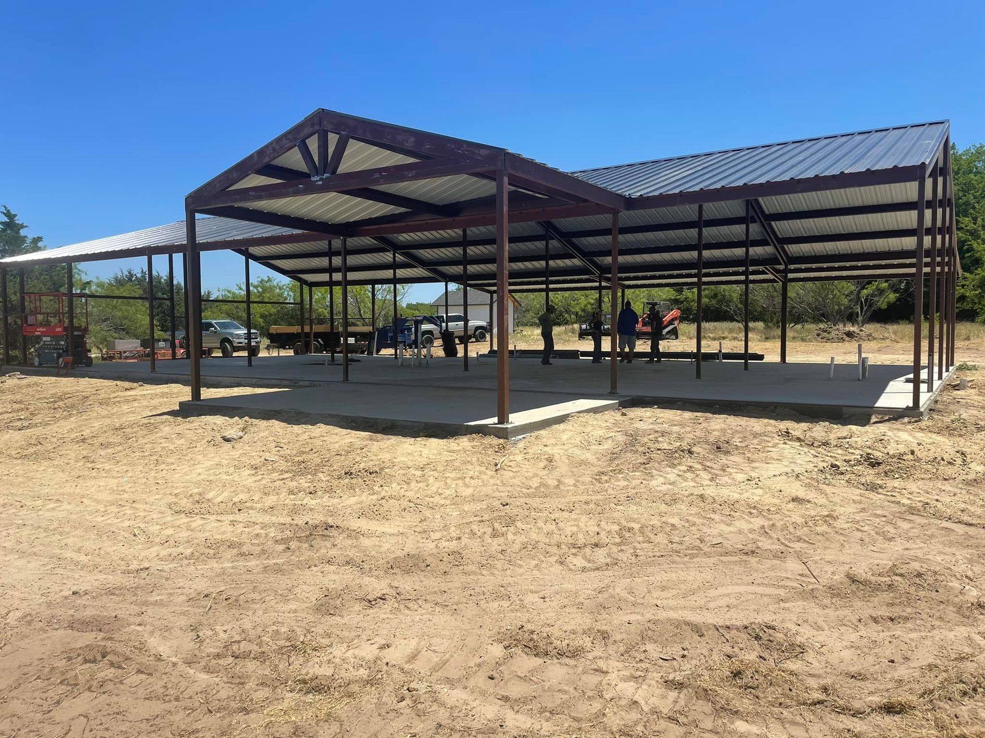 Open-air metal pavilion under construction on a sandy lot under a blue sky