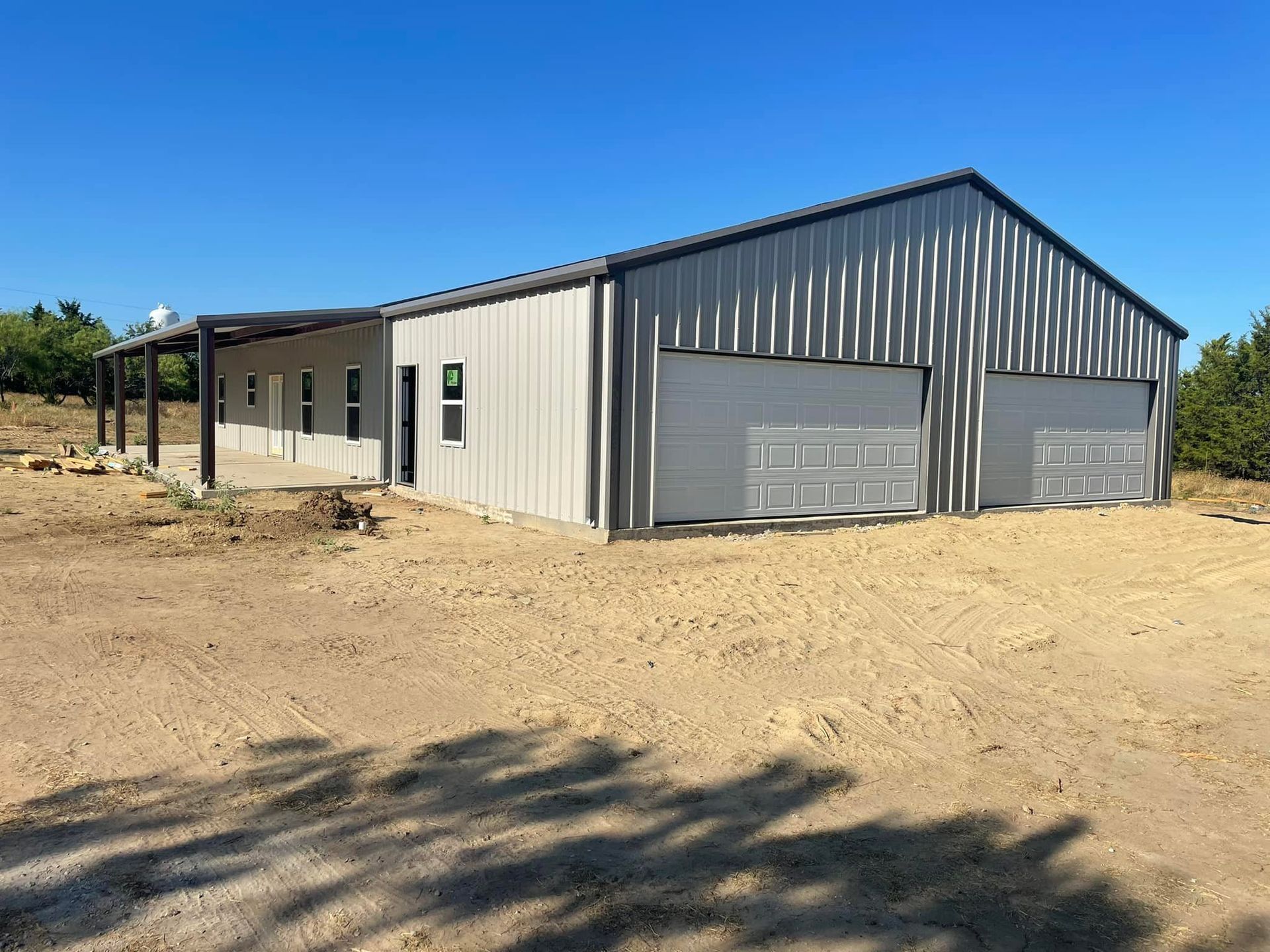 Gray metal building under construction on a dirt lot, with a gabled roof and blue sky.