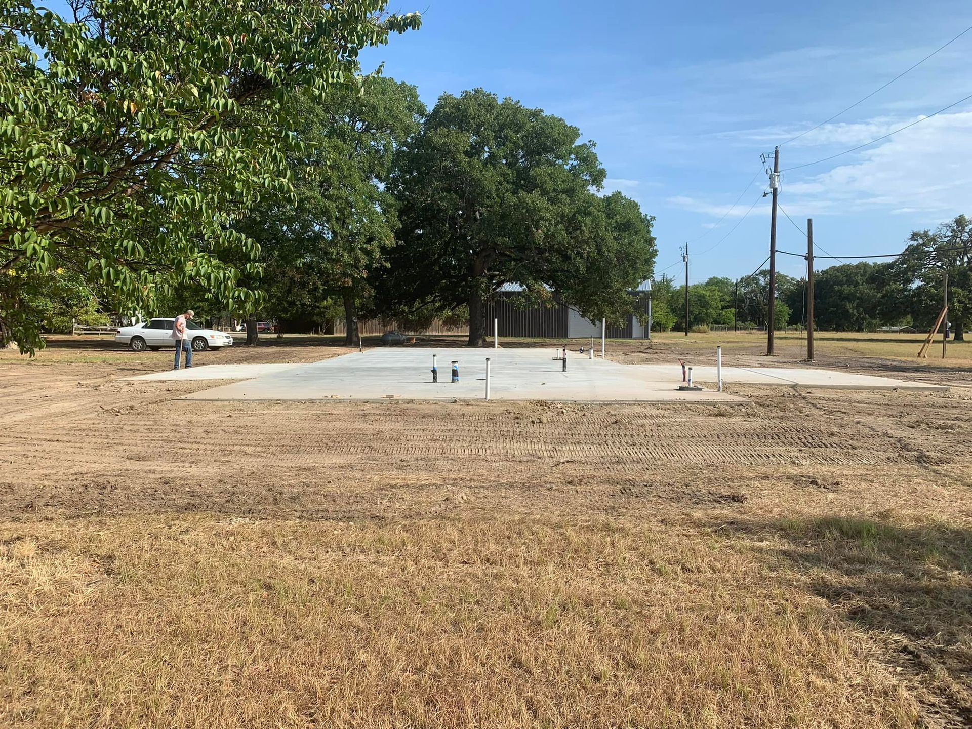 Empty concrete pad in a dry grassy lot with trees, utility poles, and a blue sky.