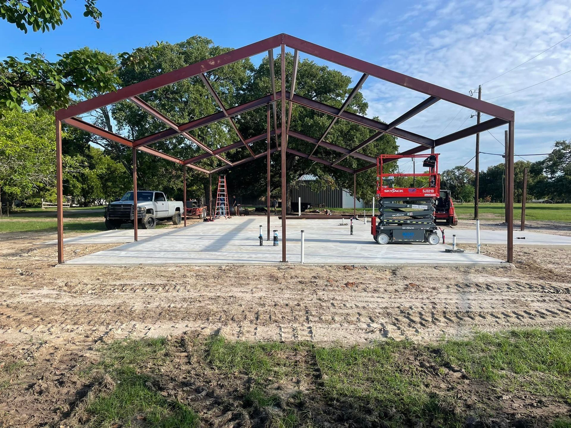 Steel building frame under construction with a concrete slab and a red lift on site.
