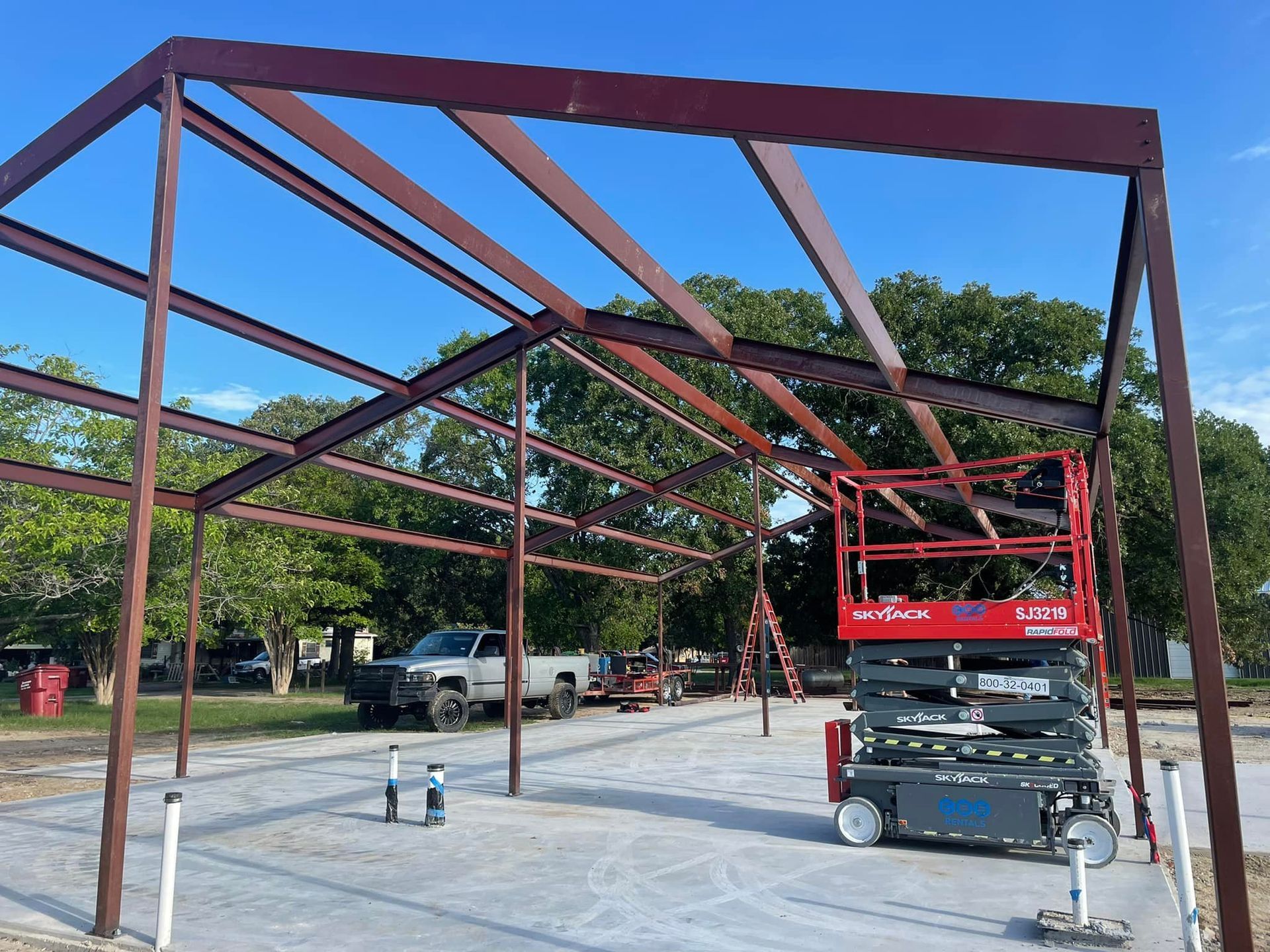 Steel building frame under construction on a concrete slab, with a red scissor lift inside.