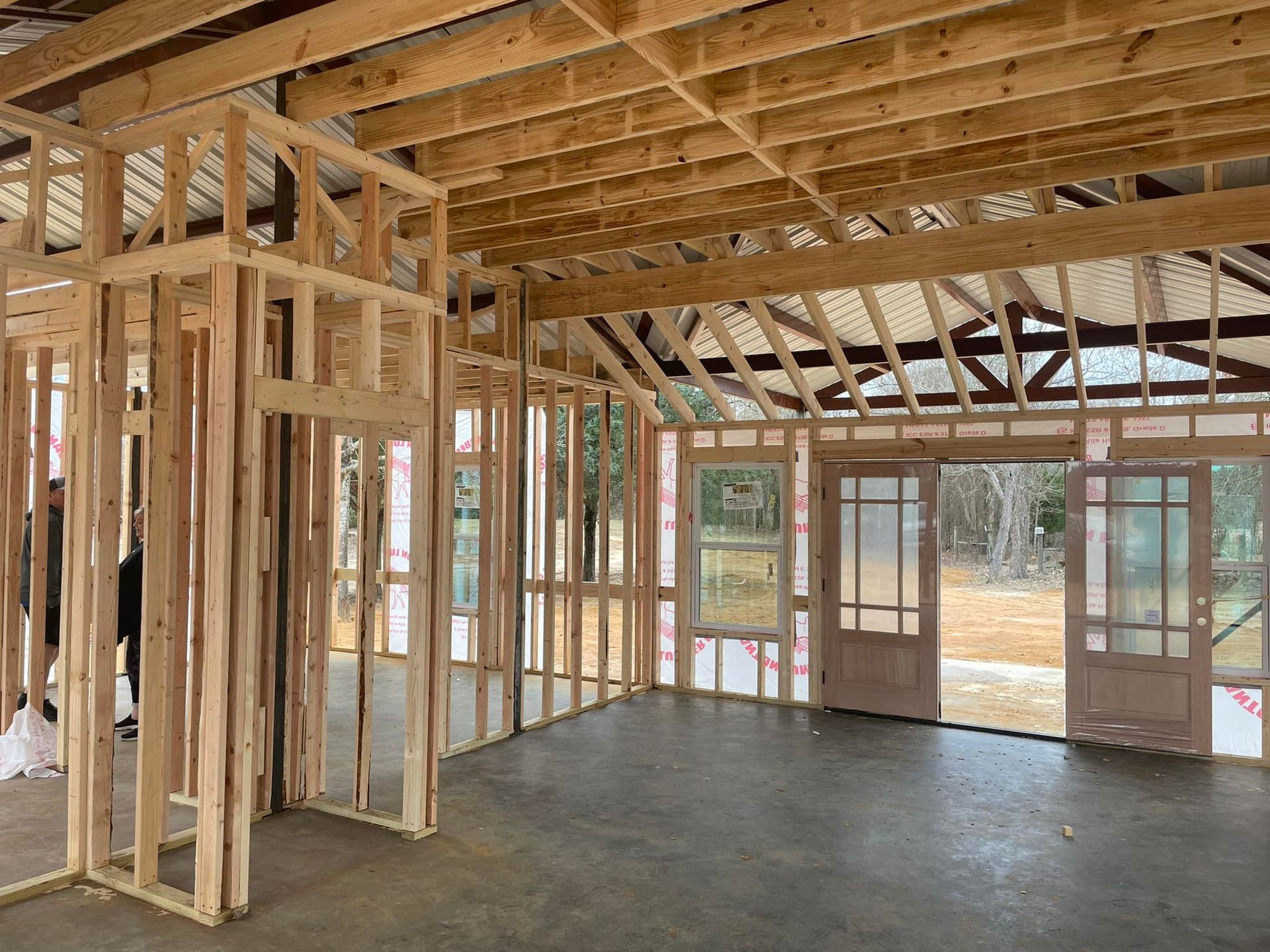 Unfinished house interior with exposed wooden framing, concrete floor, and large windows overlooking outdoors