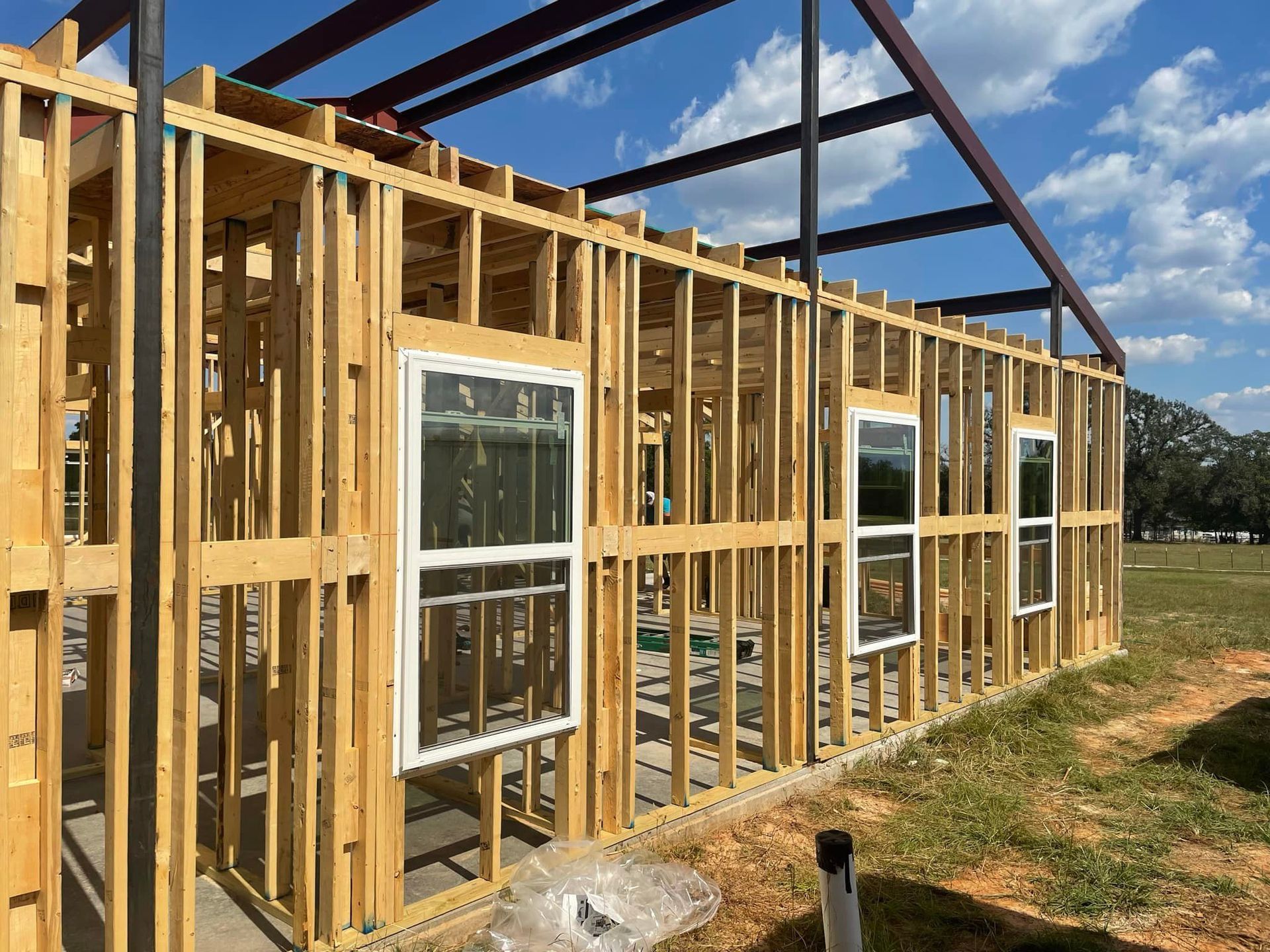 Wood-framed house under construction with exposed studs, windows, and roof trusses on a sunny lot