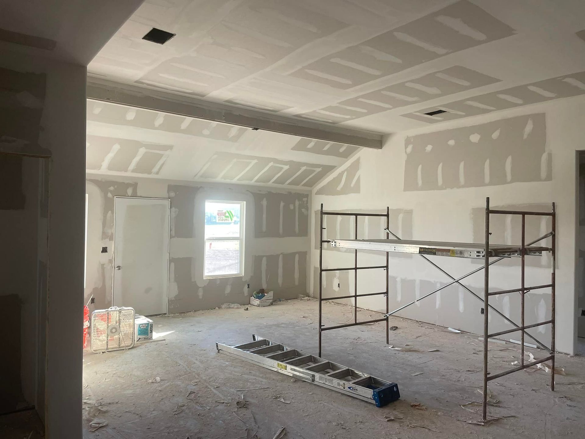 Unfinished room with drywall patches, scaffolding, debris, and a window letting in daylight