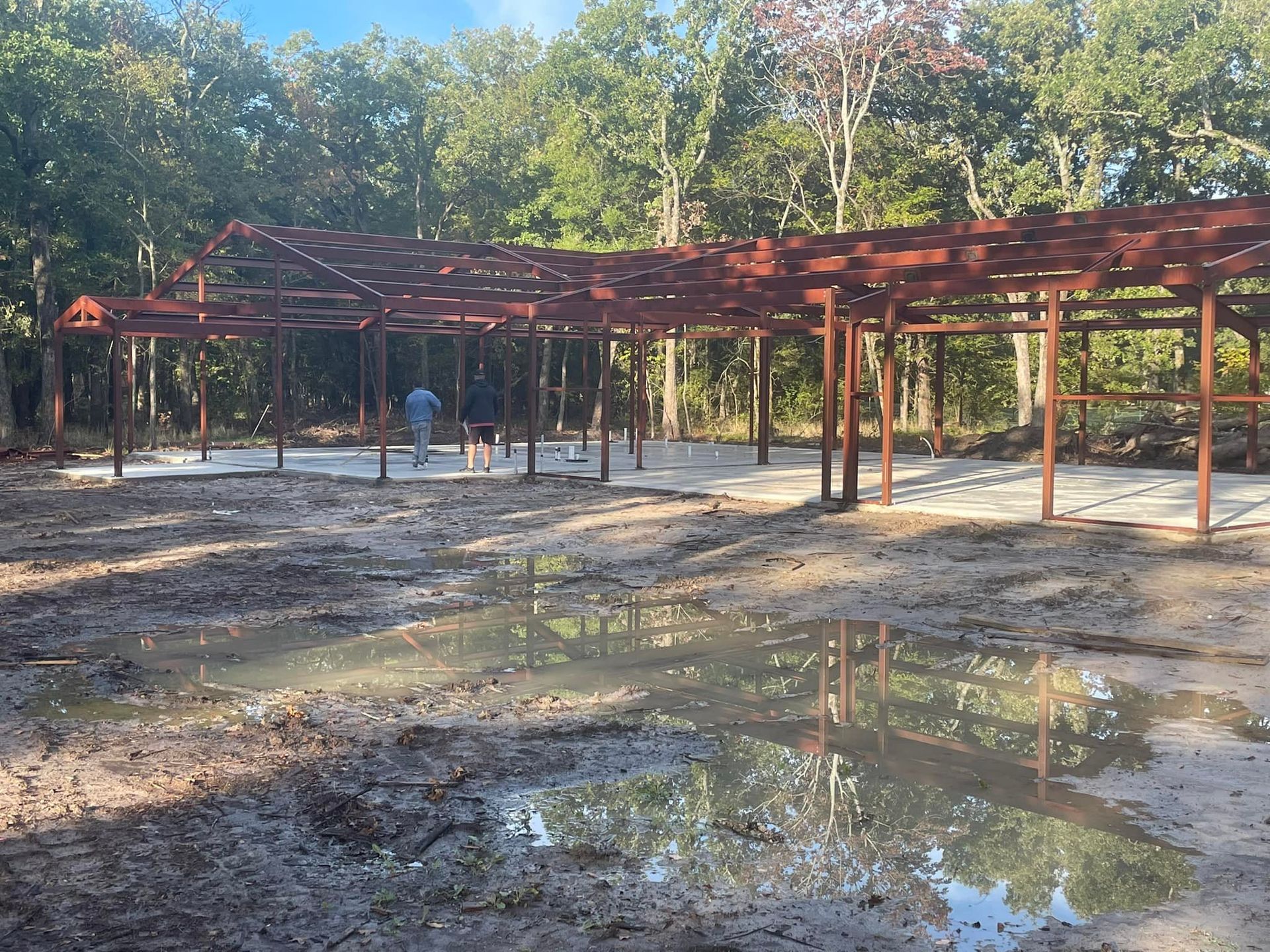 Rusty metal pavilion frame on a muddy, puddled lot with trees in the background