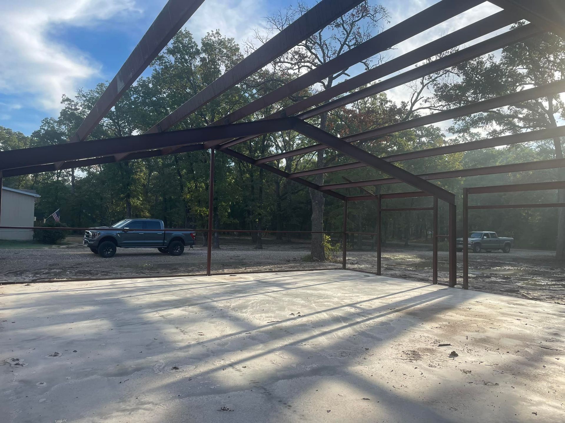 Open steel carport frame casting long shadows on a concrete slab, with trucks parked nearby and trees in back.