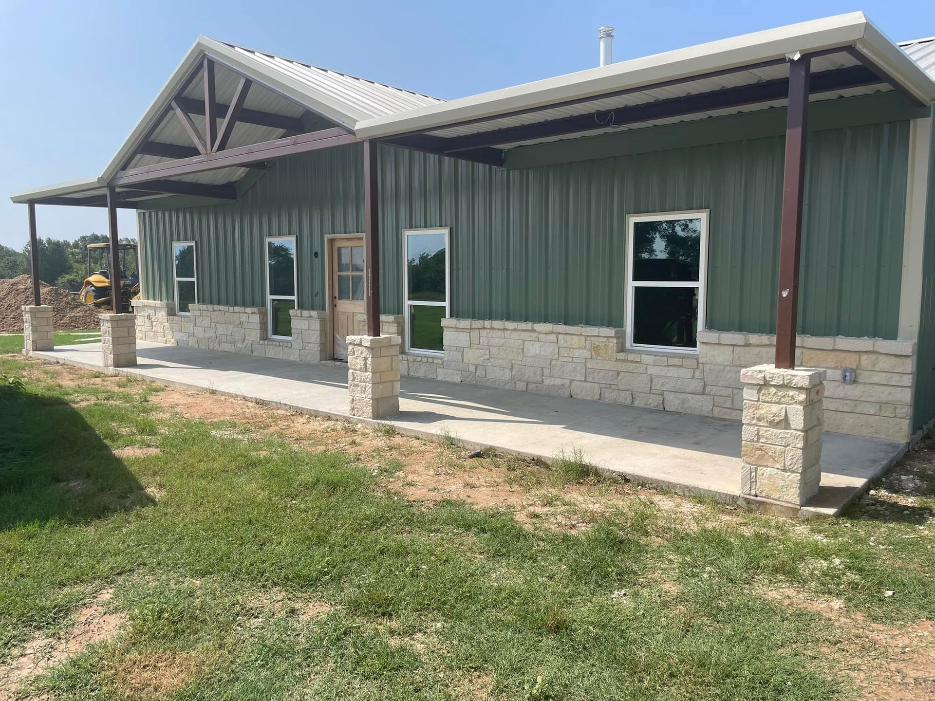Small green metal building with stone columns and a covered porch on a grassy lot.