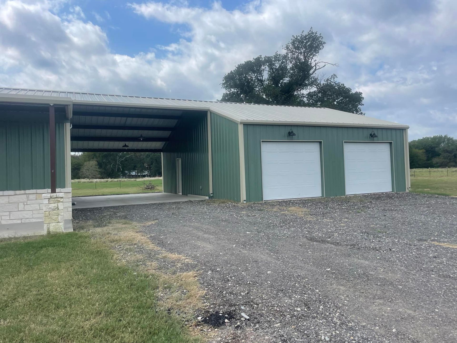 Green metal garage with two white doors beside an open bay on a gravel driveway under a cloudy sky