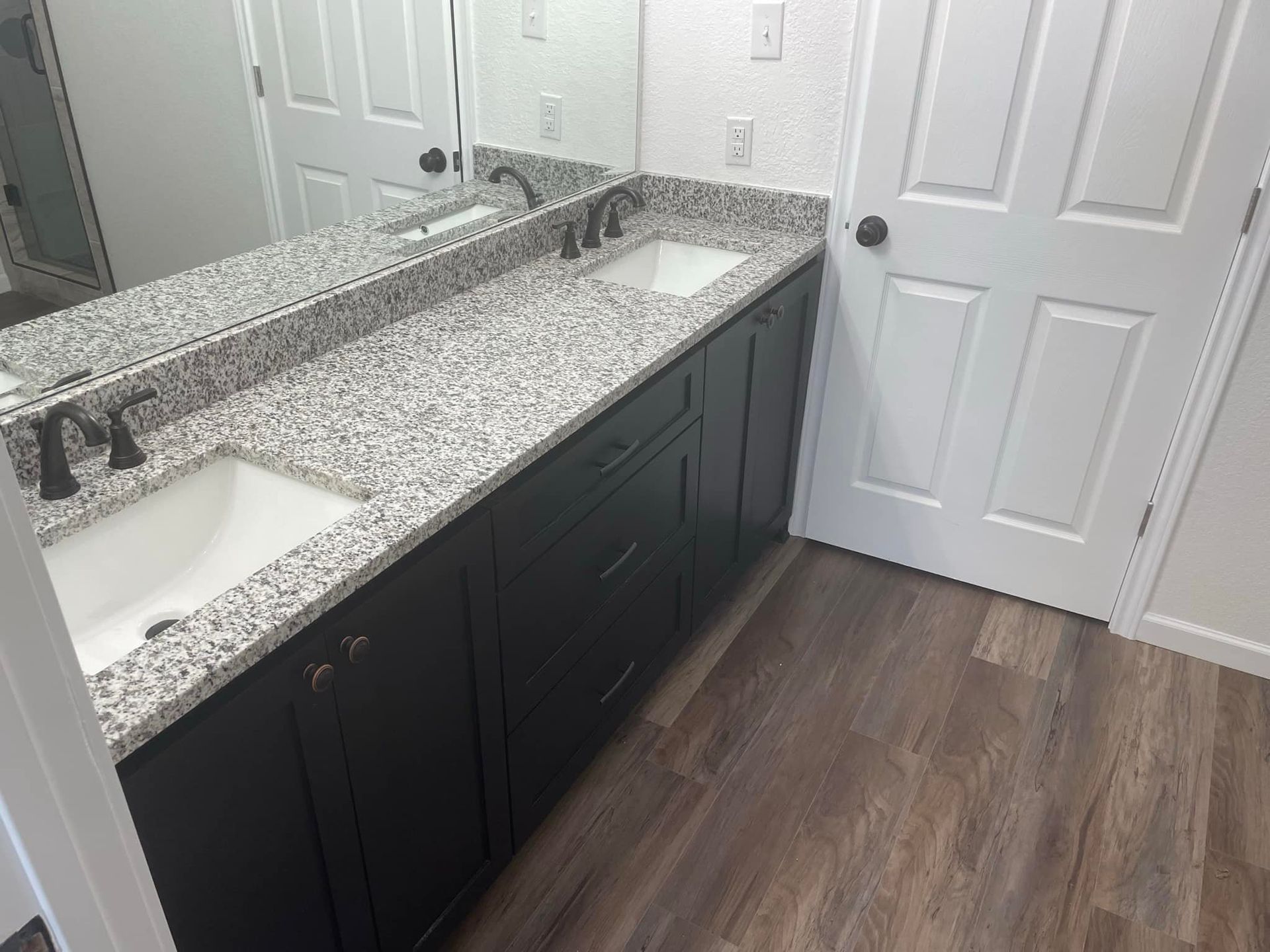 Bathroom vanity with double sinks, speckled granite countertop, black cabinets, and wood-look flooring