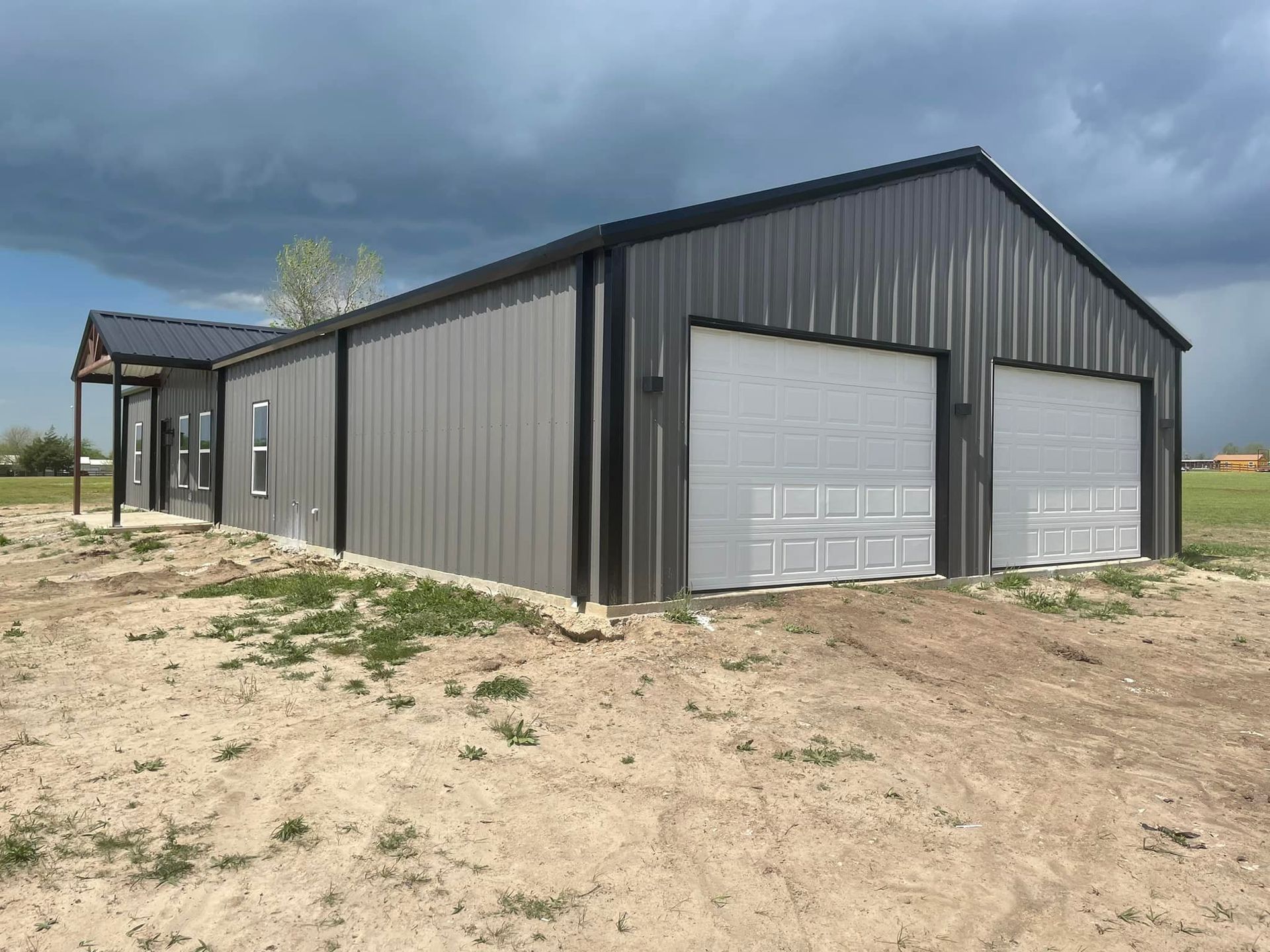 Gray metal barn with white garage doors on a dirt lot under a cloudy sky