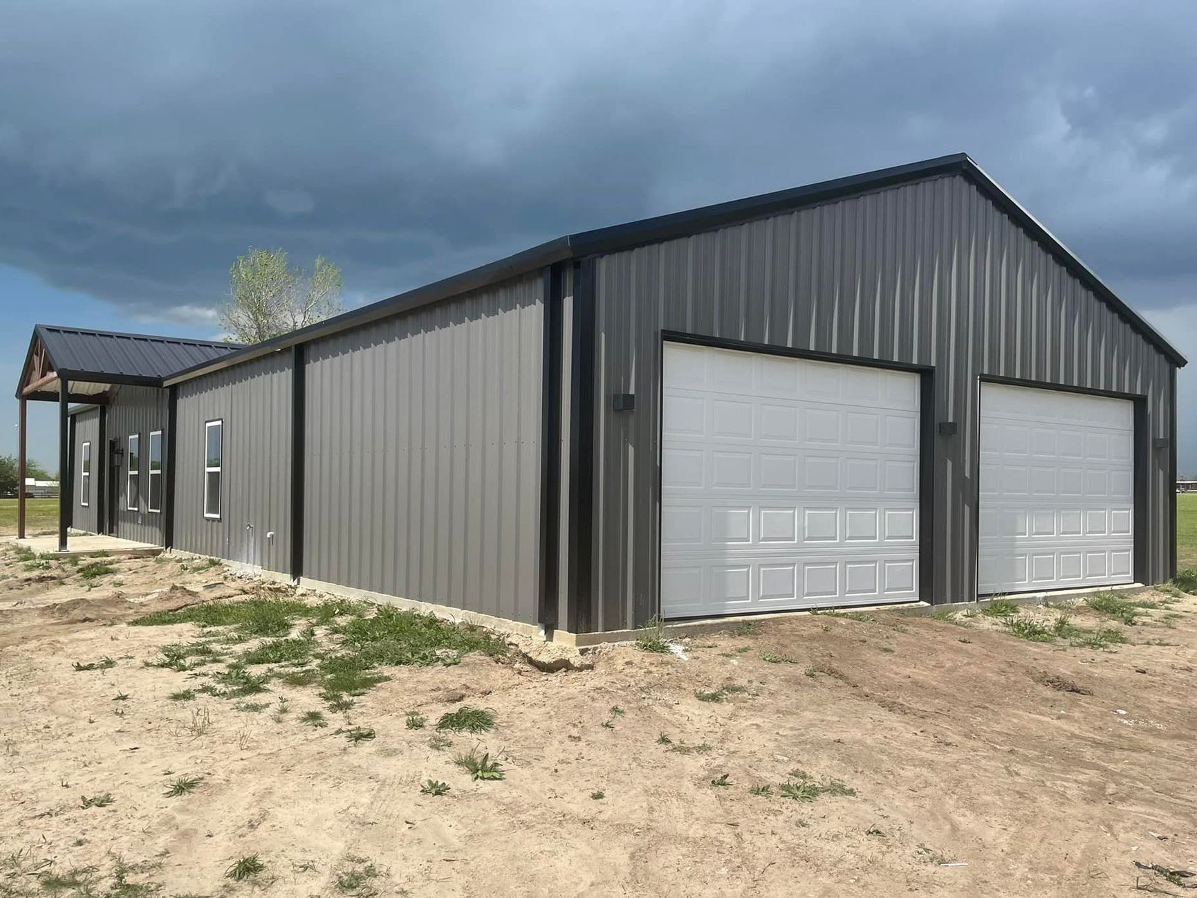 Gray metal barn with two garage doors on a dirt lot under a cloudy sky