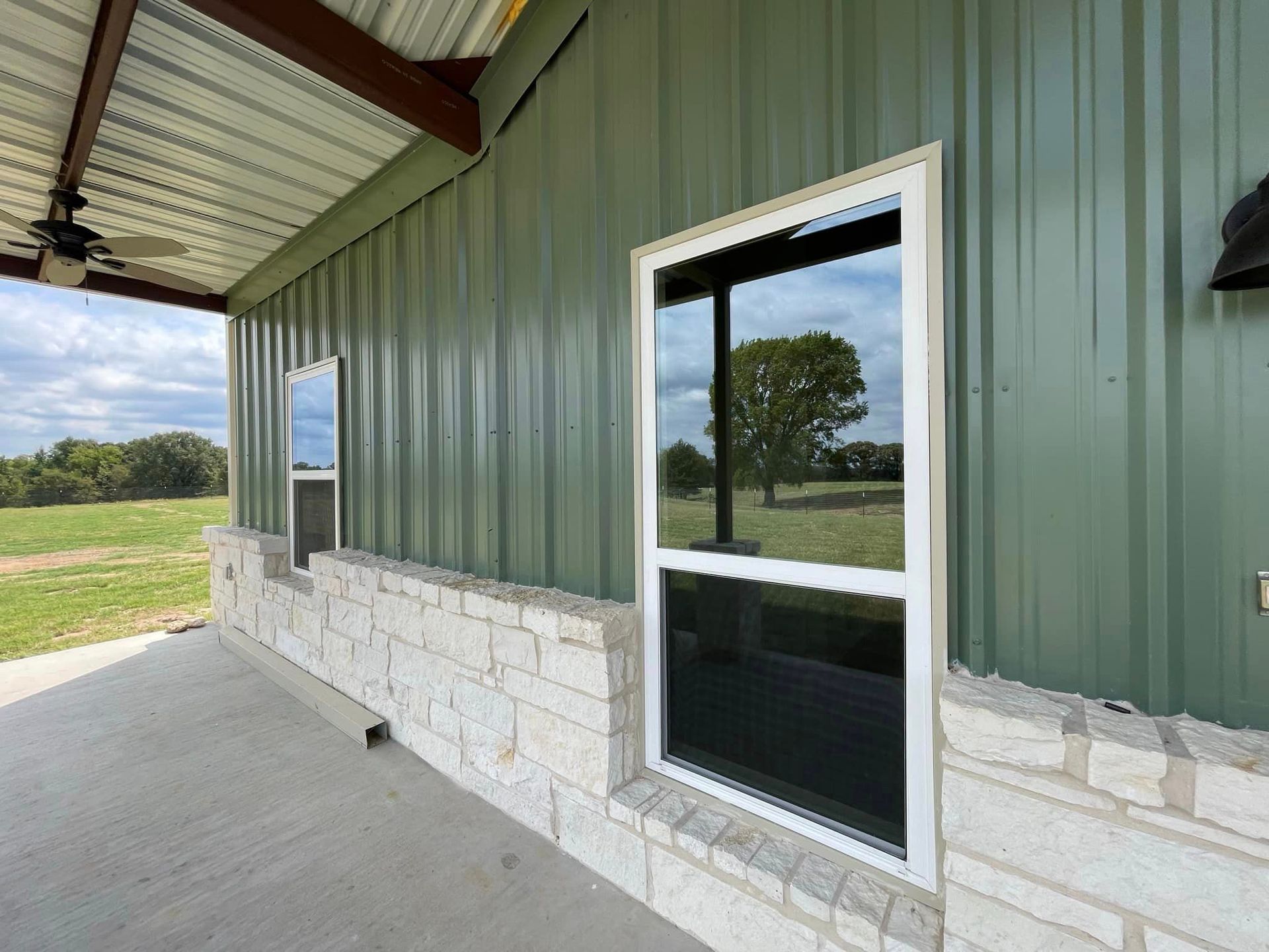 Covered porch with green metal siding, stone base, and a glass door overlooking a grassy field.