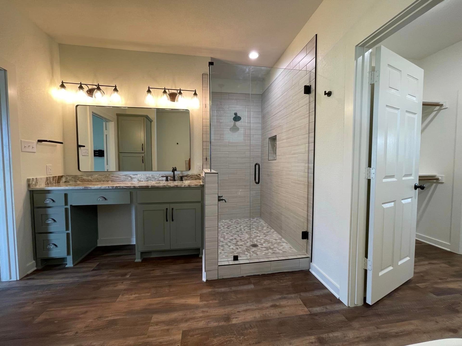 Bathroom with double vanity, large mirror, tiled shower, and open white door on wood floor.