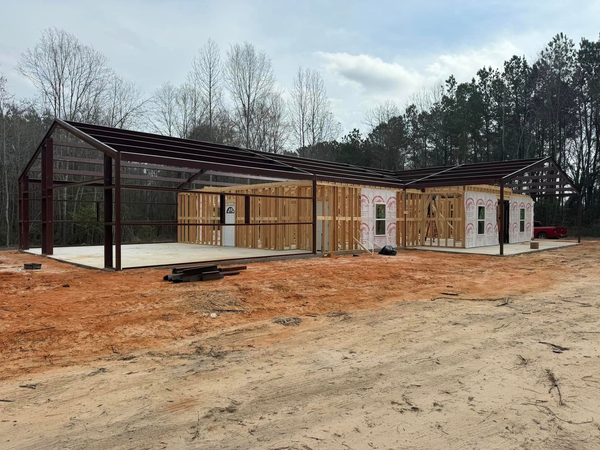 Partially built wooden house frame with metal roof trusses on a dirt lot under a cloudy sky
