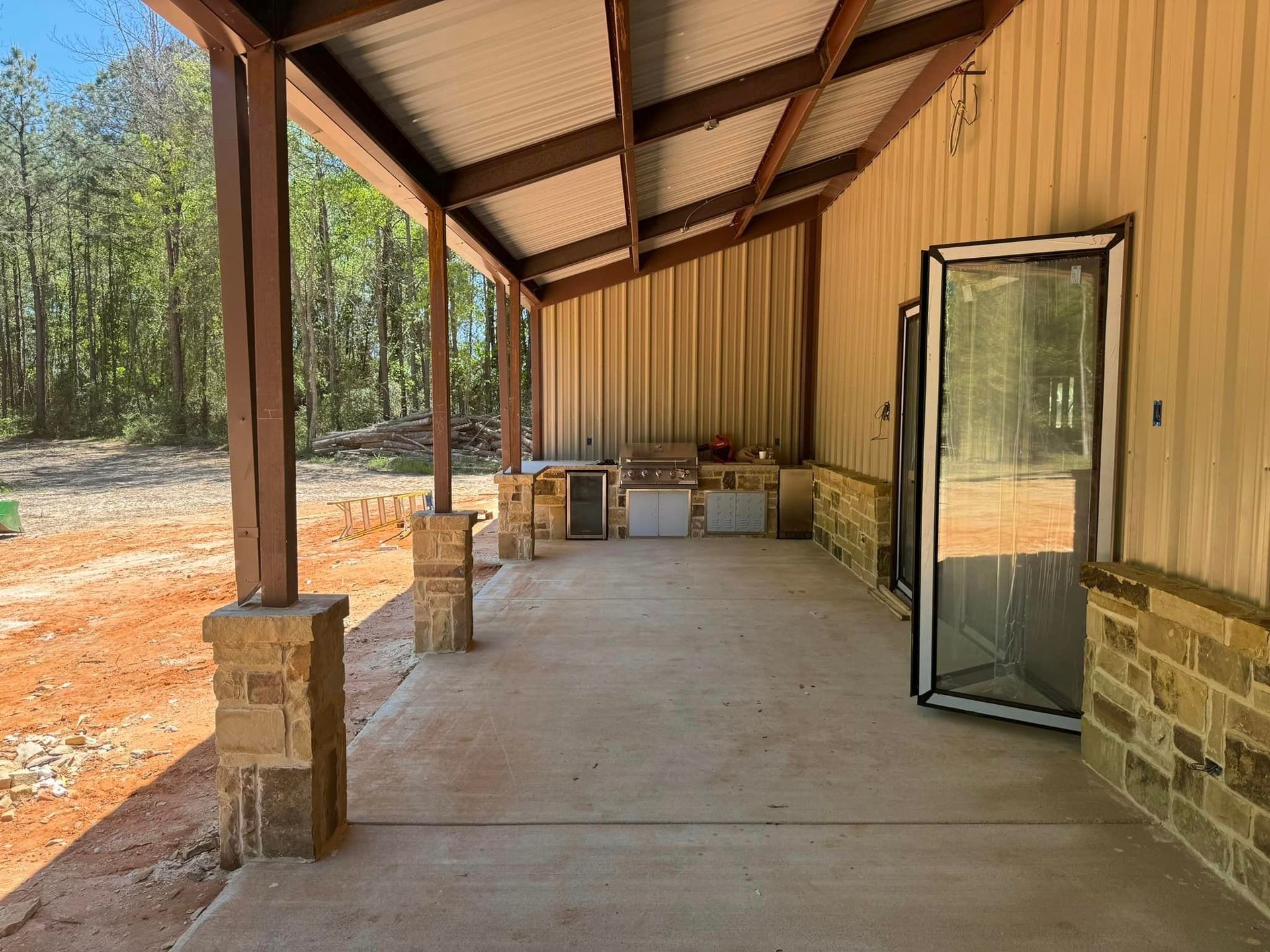 Covered porch with concrete floor, tan metal walls, and an open glass door beside a wooded yard.