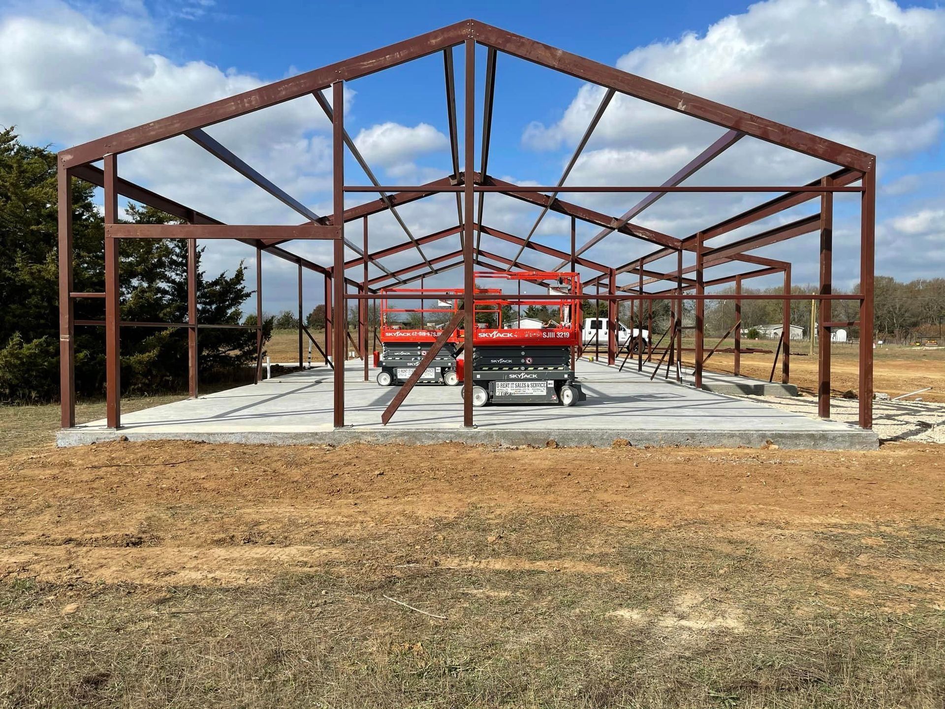 Steel building frame under construction with a red lift on a concrete slab, viewed across a dirt lot.