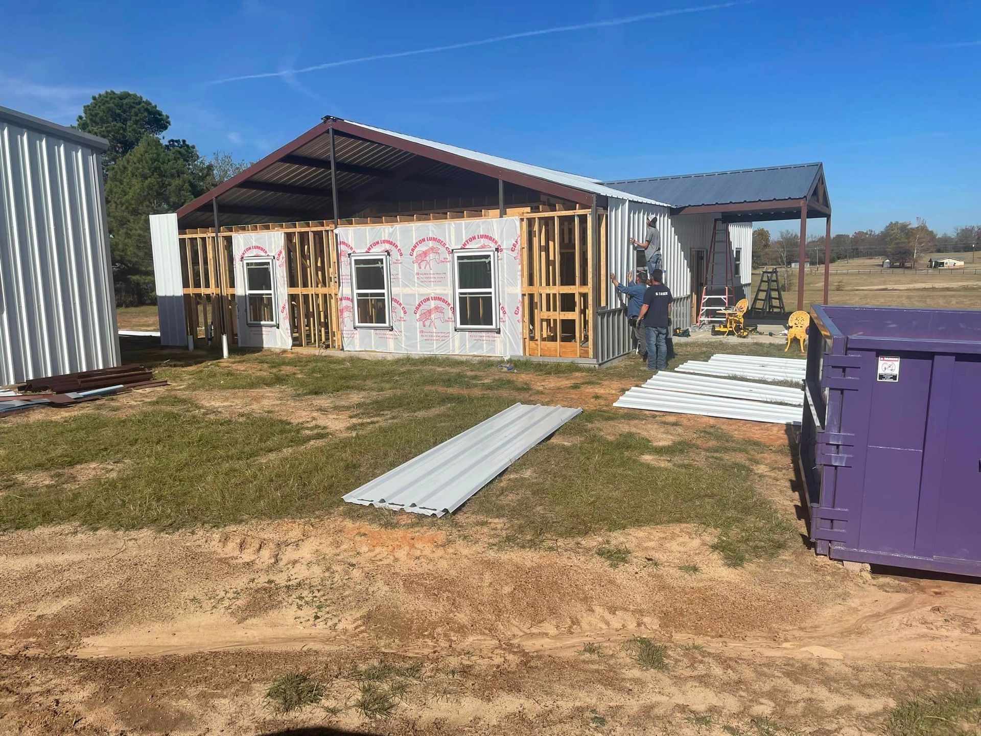 Partially built house with exposed framing, metal sheets, and a purple dumpster in the yard