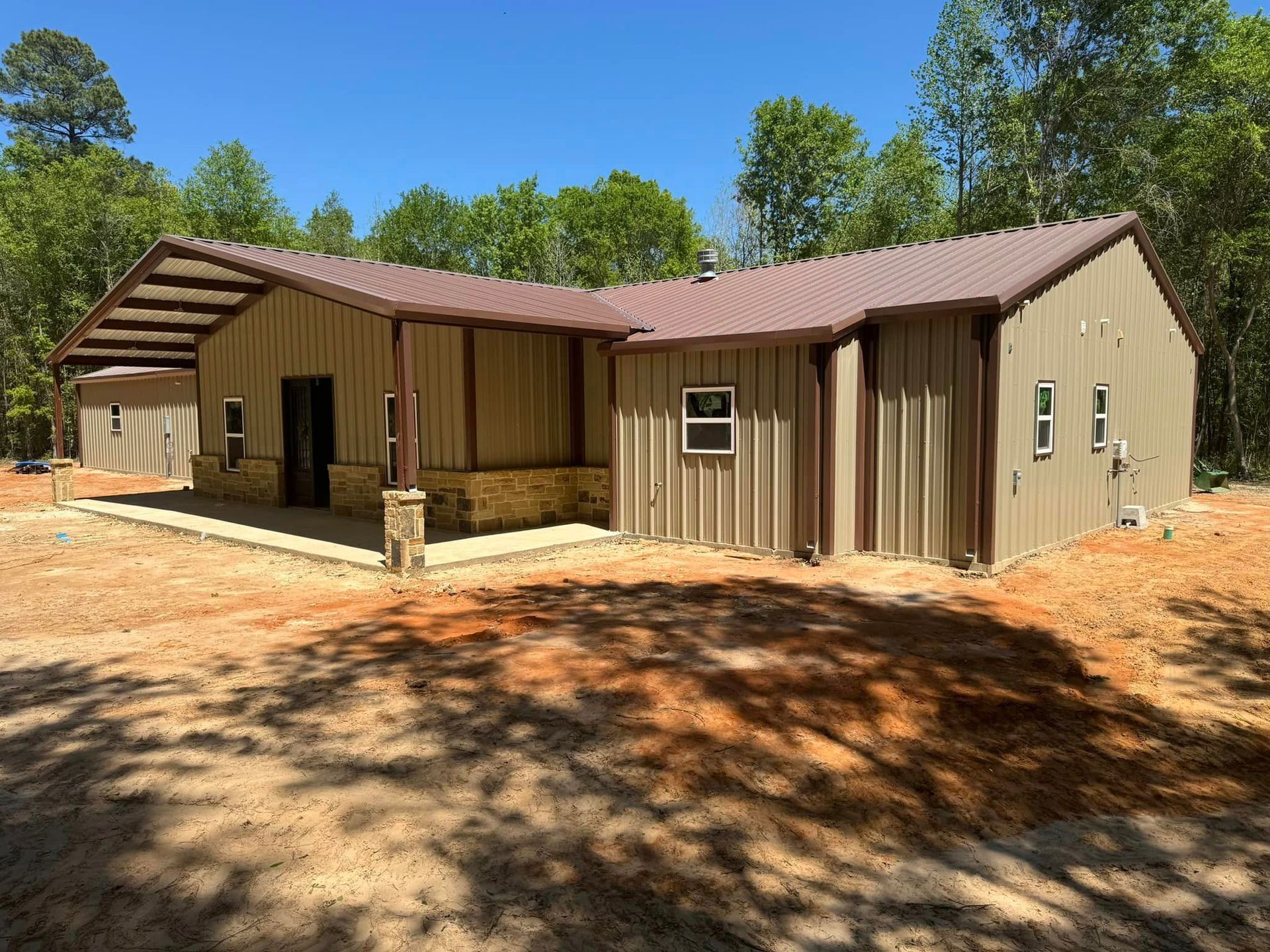 Single-story tan cabin with porch and carport in a wooded clearing under a blue sky