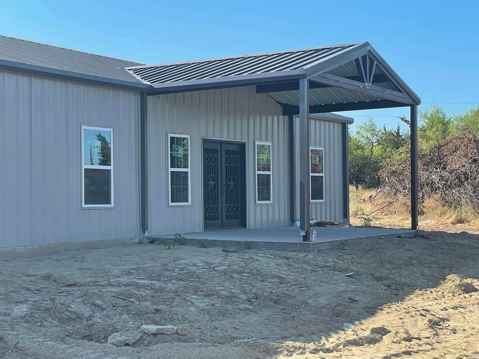 Gray building with a covered entry, metal roof, and dirt foreground under a clear blue sky