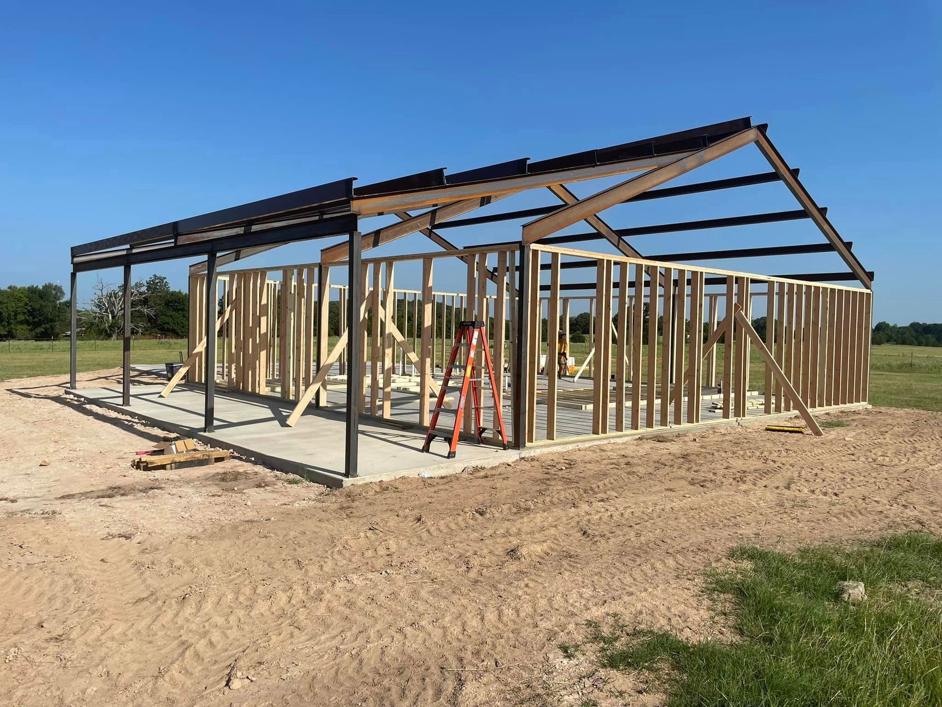 Wood-framed house under construction with steel roof beams, a ladder, and open dirt lot under a blue sky