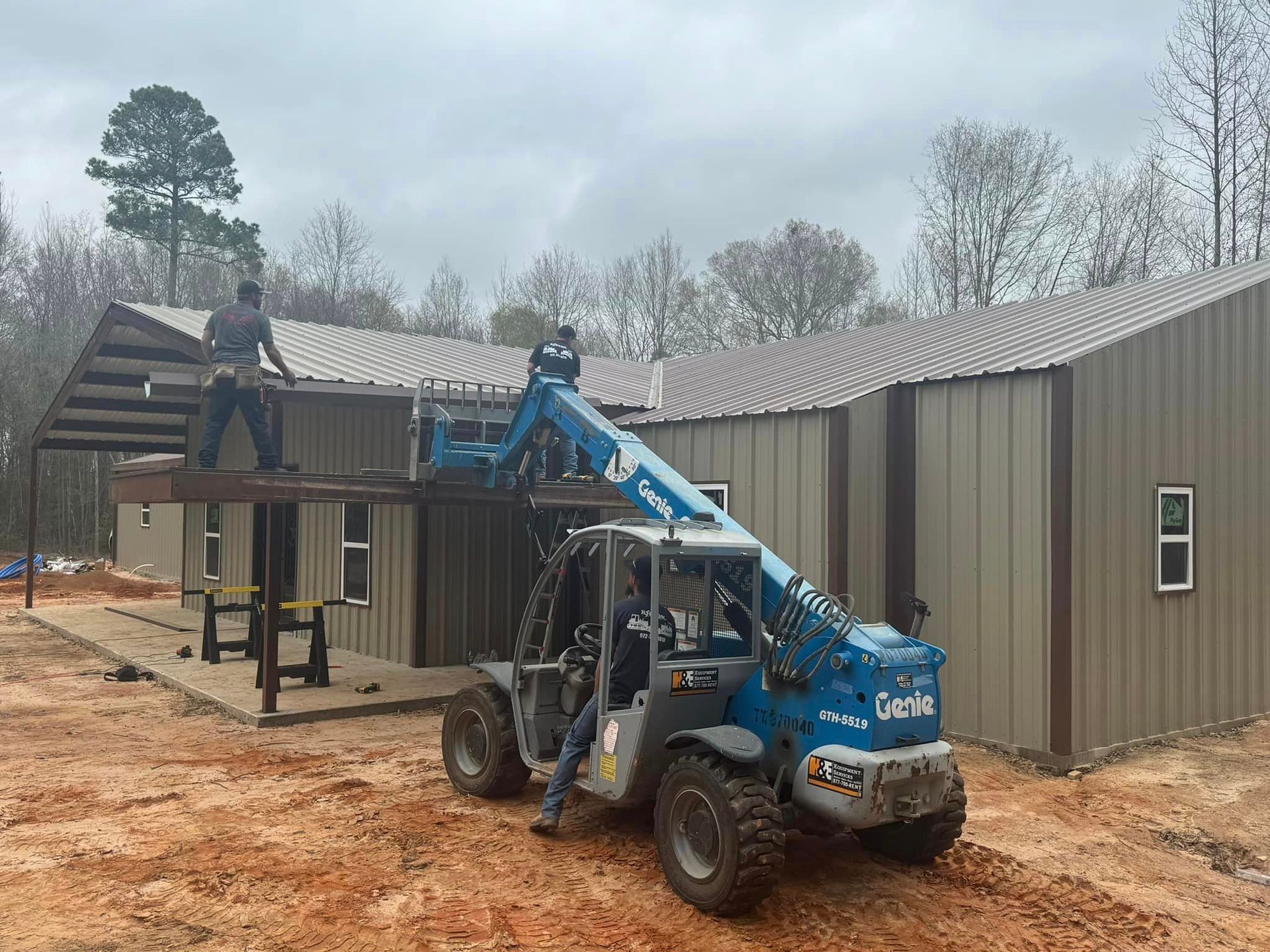 Workers install metal siding on a house using a blue forklift on a muddy construction site.