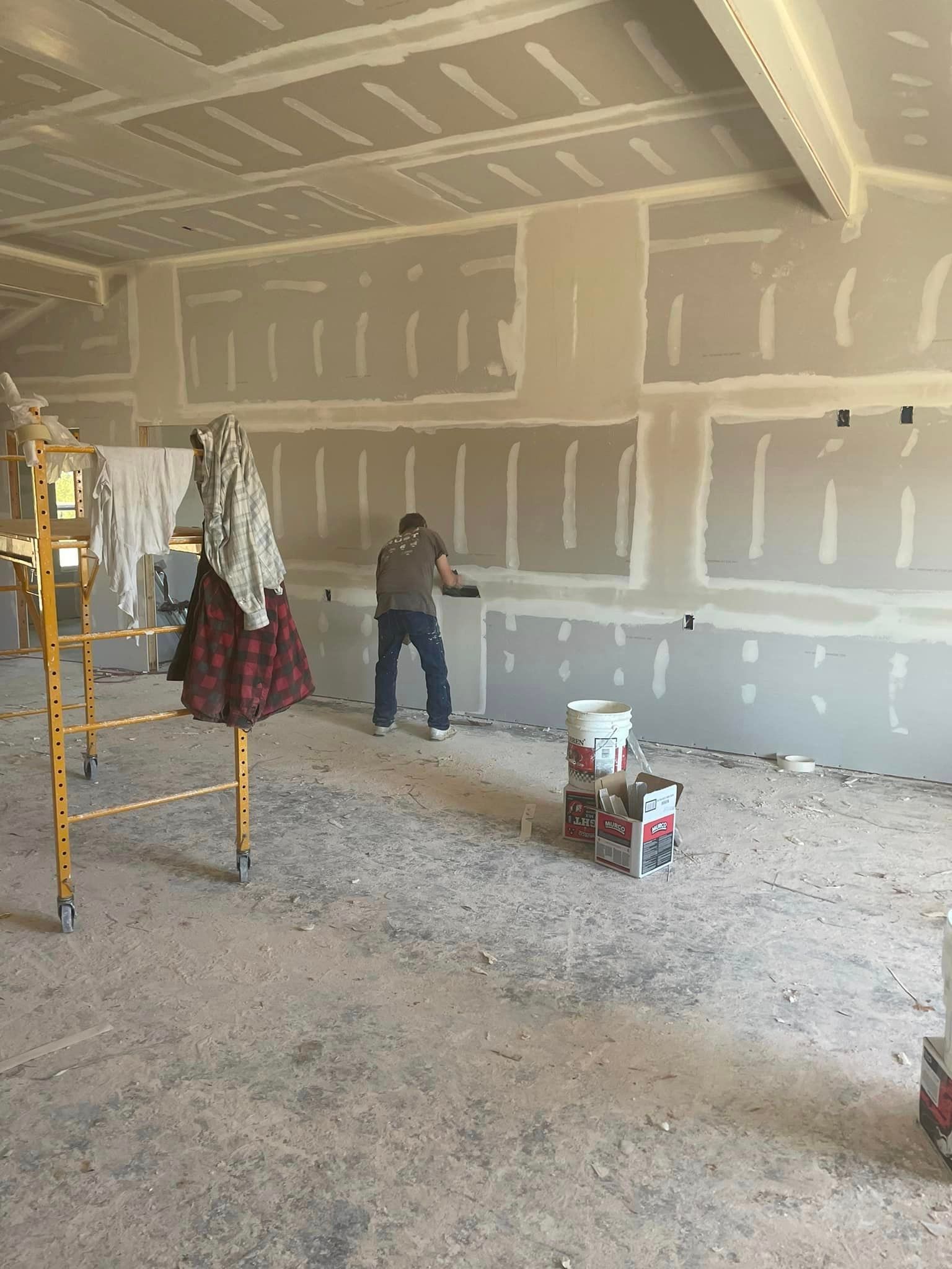 Unfinished drywall room with a person standing near the back wall and tools on a scaffold in the foreground