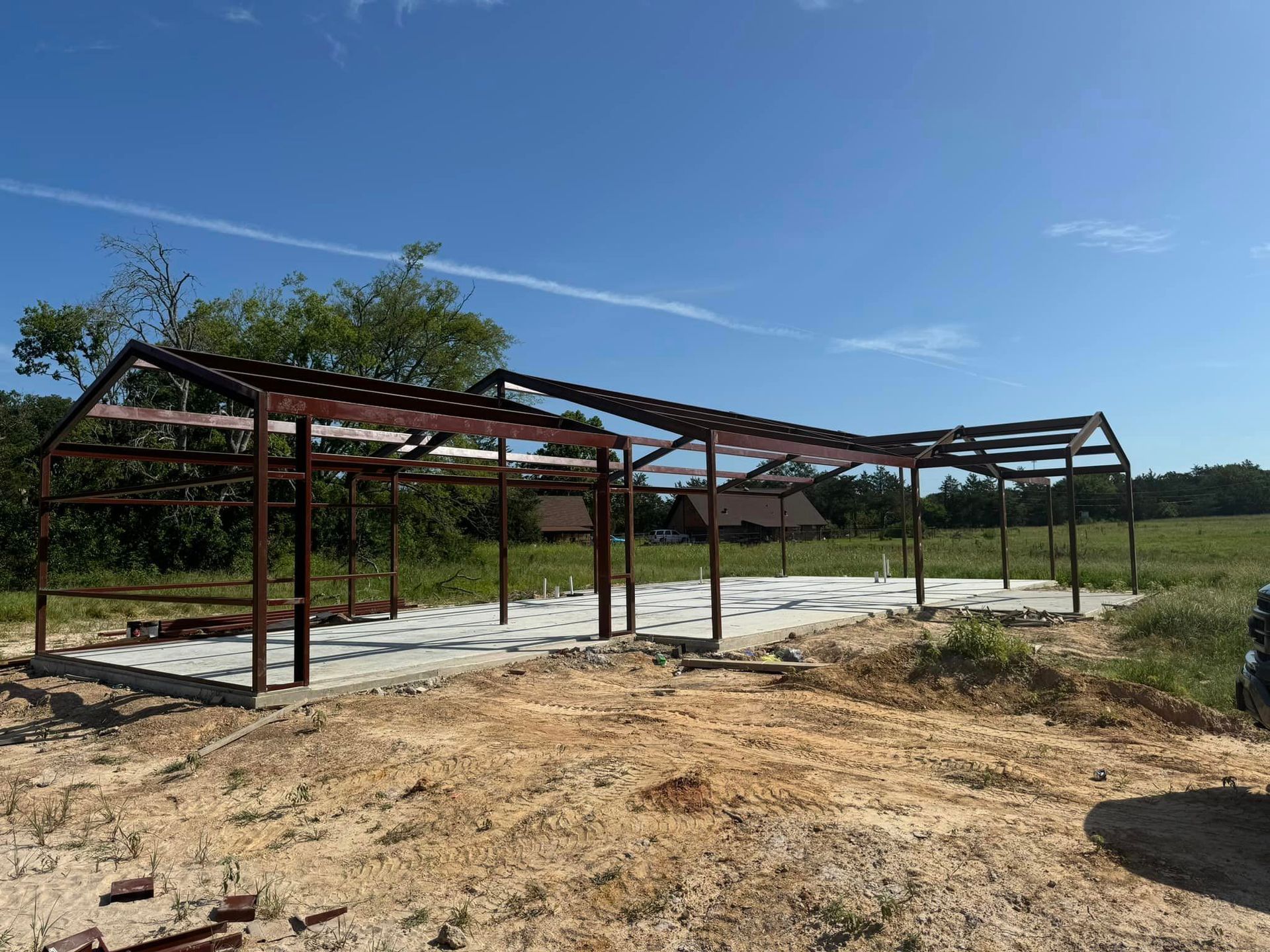 Partially built steel structure on a concrete slab in a rural field under a clear blue sky
