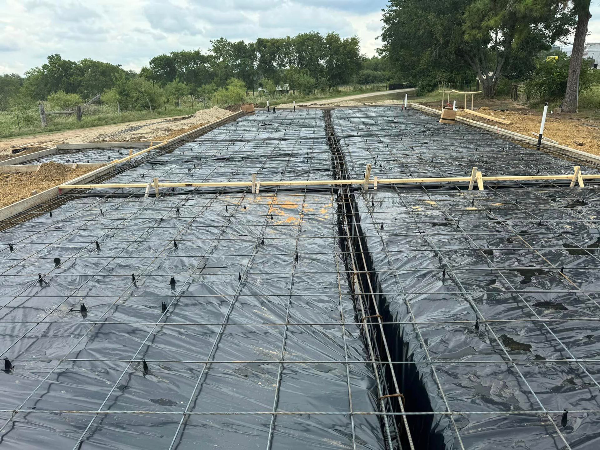 Concrete slab under construction with black plastic sheeting, rebar stakes, and wooden forms along a road.