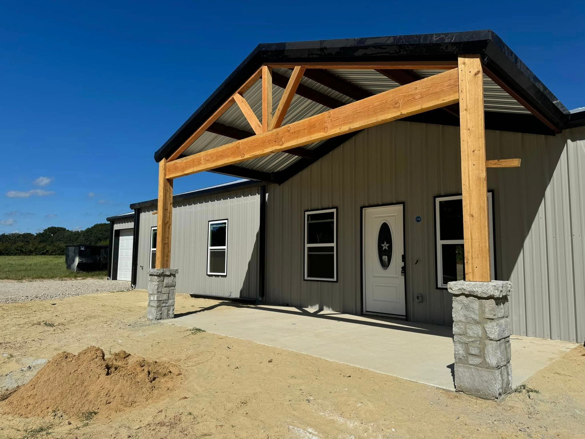Modern single-story building with wood truss porch and stone columns on a dirt lot under blue sky