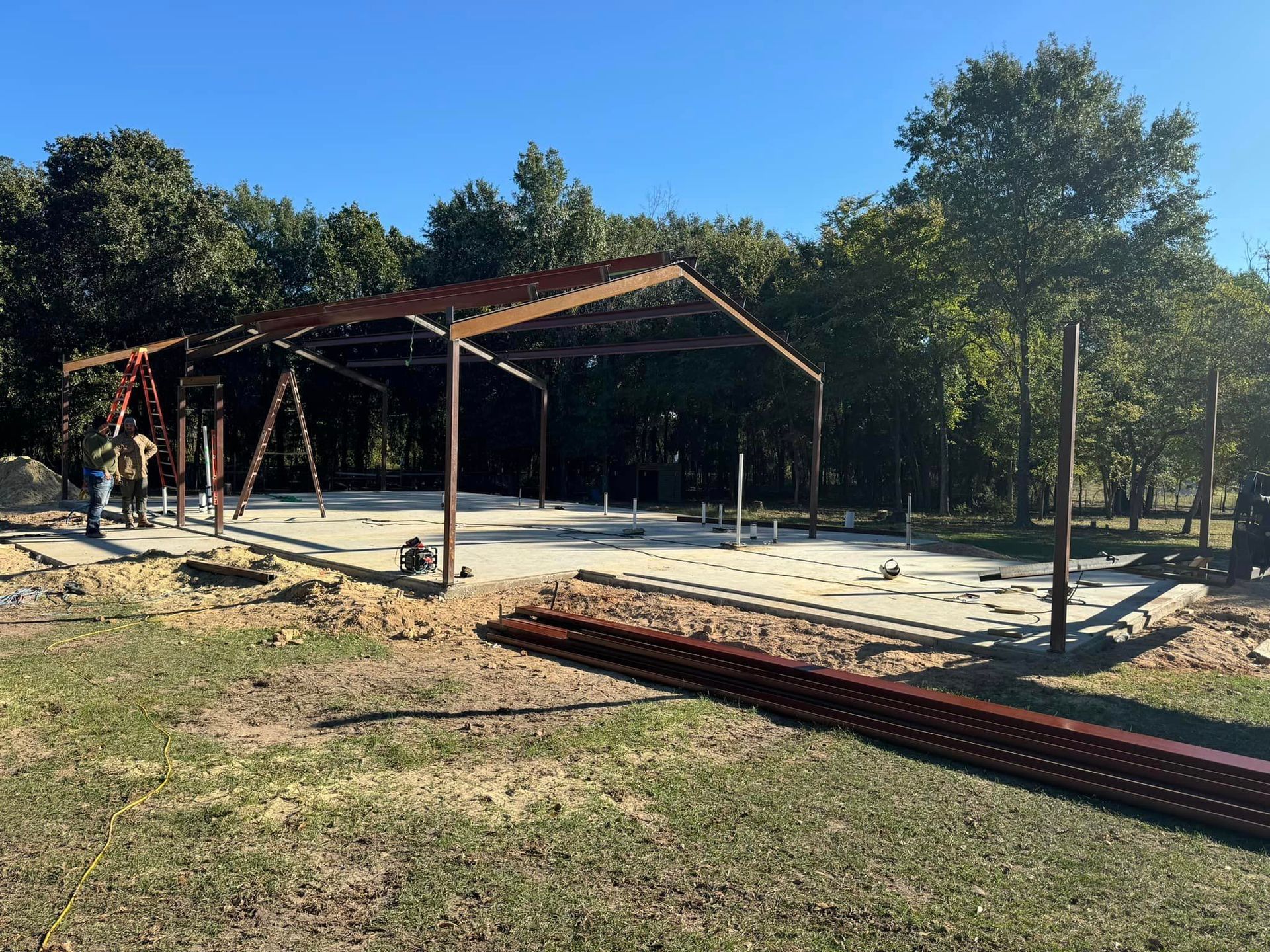 Partially built wooden pavilion on a concrete slab in a sunny, wooded park area