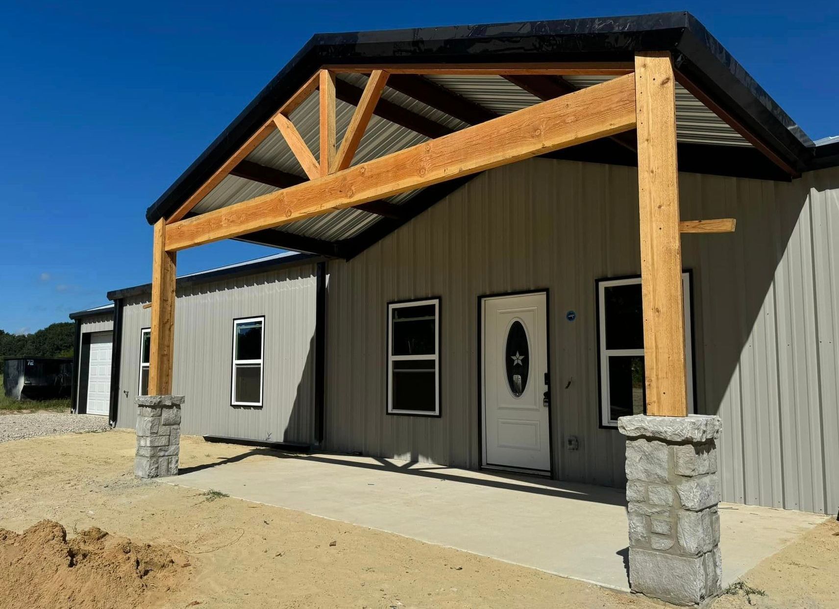 A small gray building with a wooden porch roof and stone columns under a clear blue sky.