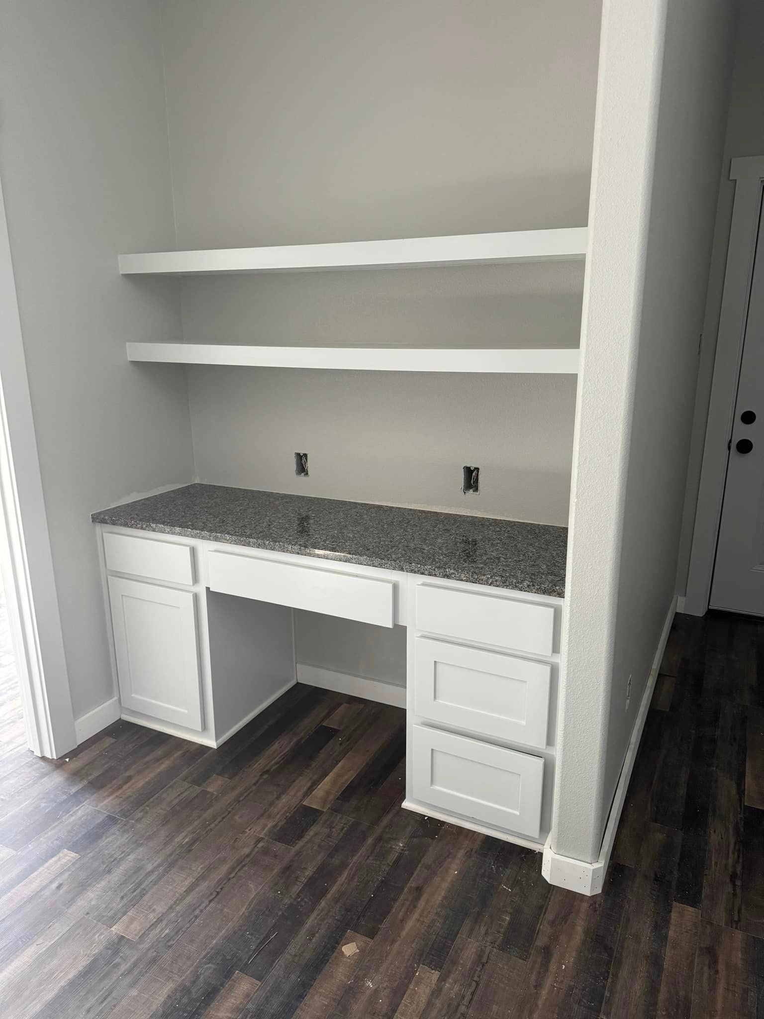 Built-in desk nook with gray countertop, white shelves, cabinets, and dark wood flooring