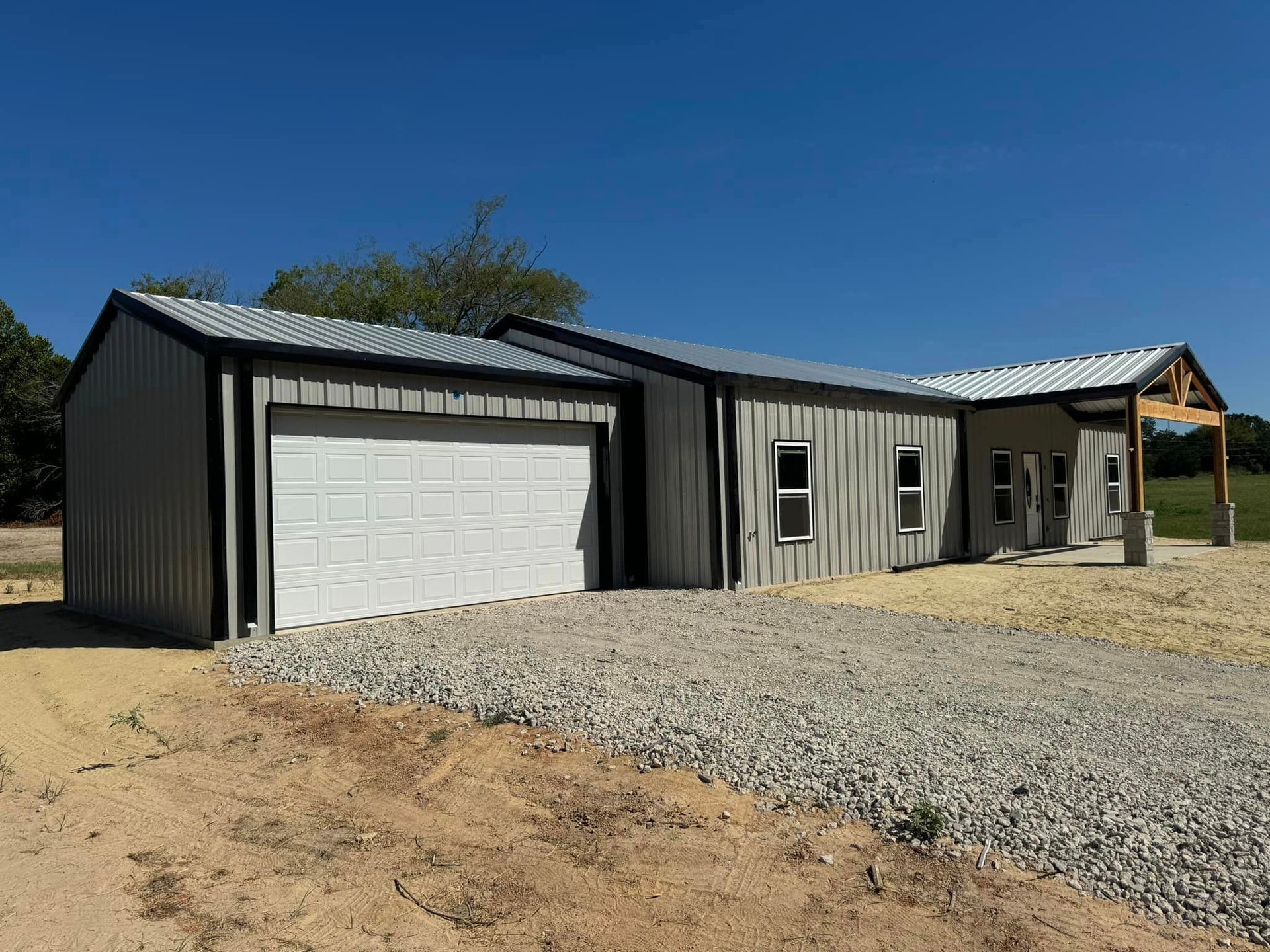 Single-story gray metal house with attached white garage and front porch on a gravel lot under a clear blue sky