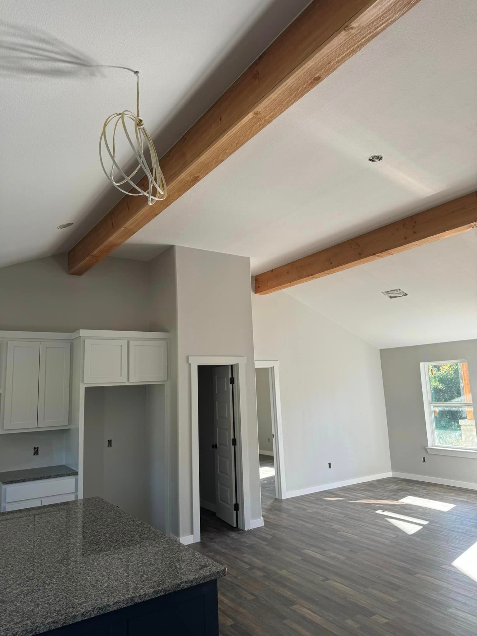 Open kitchen and living room with exposed wood beams, white walls, gray floor, and sunlight from windows.