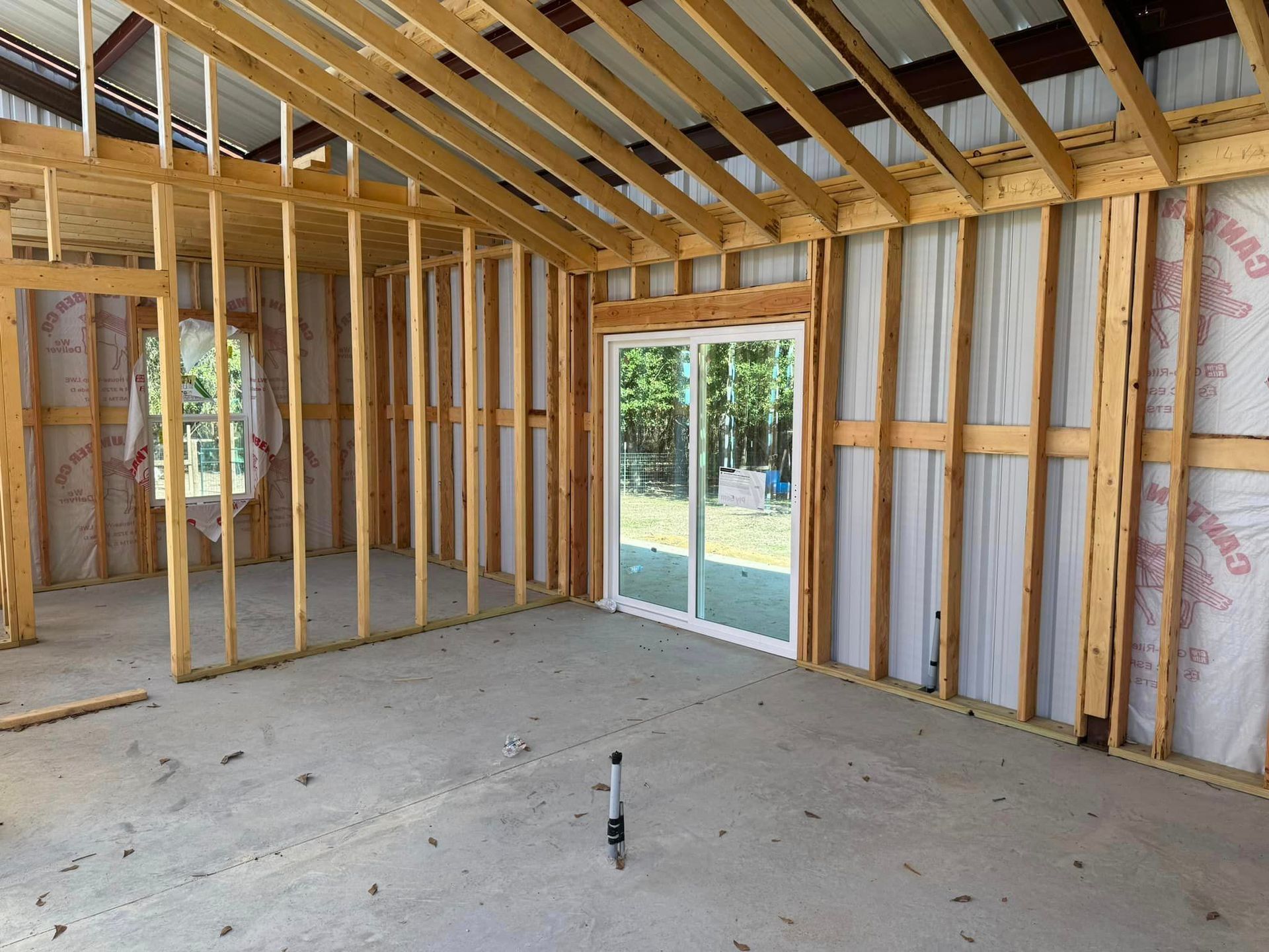 Unfinished room with exposed wooden framing, plywood floor, and a sliding glass door to a backyard.