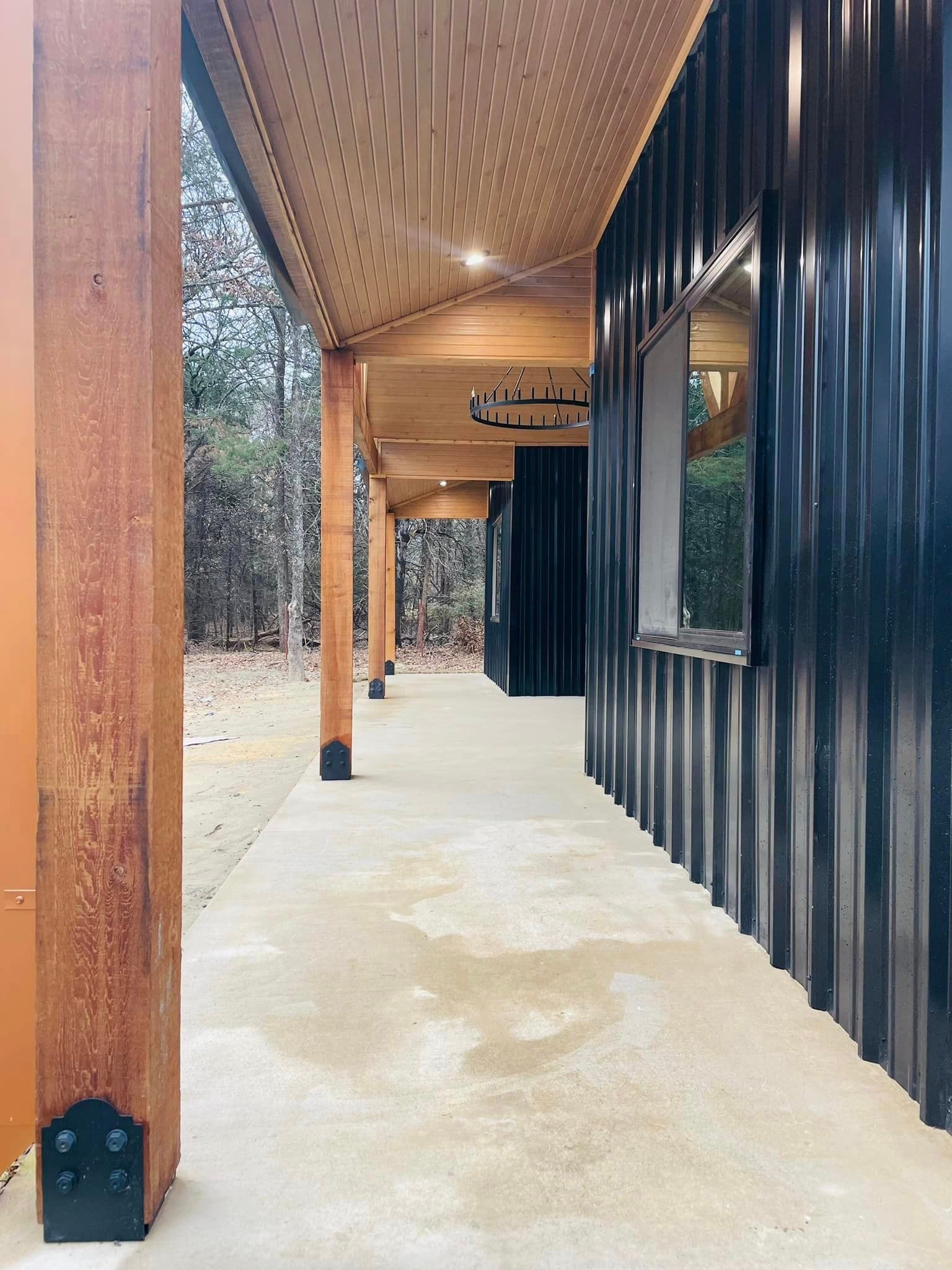 Covered walkway with wooden posts, black metal siding, and concrete floor leading toward a distant doorway