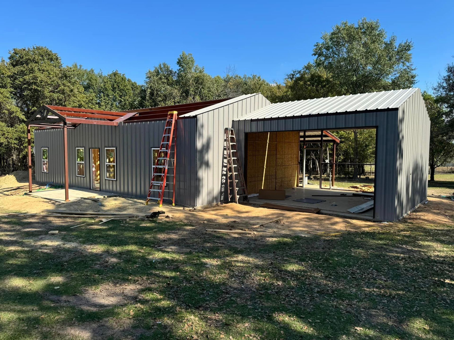Partially built modular building with metal framing and open bays on a sunny wooded lot