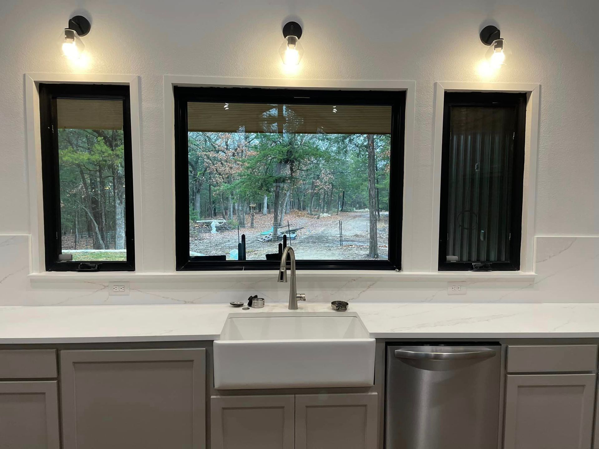 Modern kitchen sink beneath three windows, with white countertop, gray cabinets, and stainless dishwasher