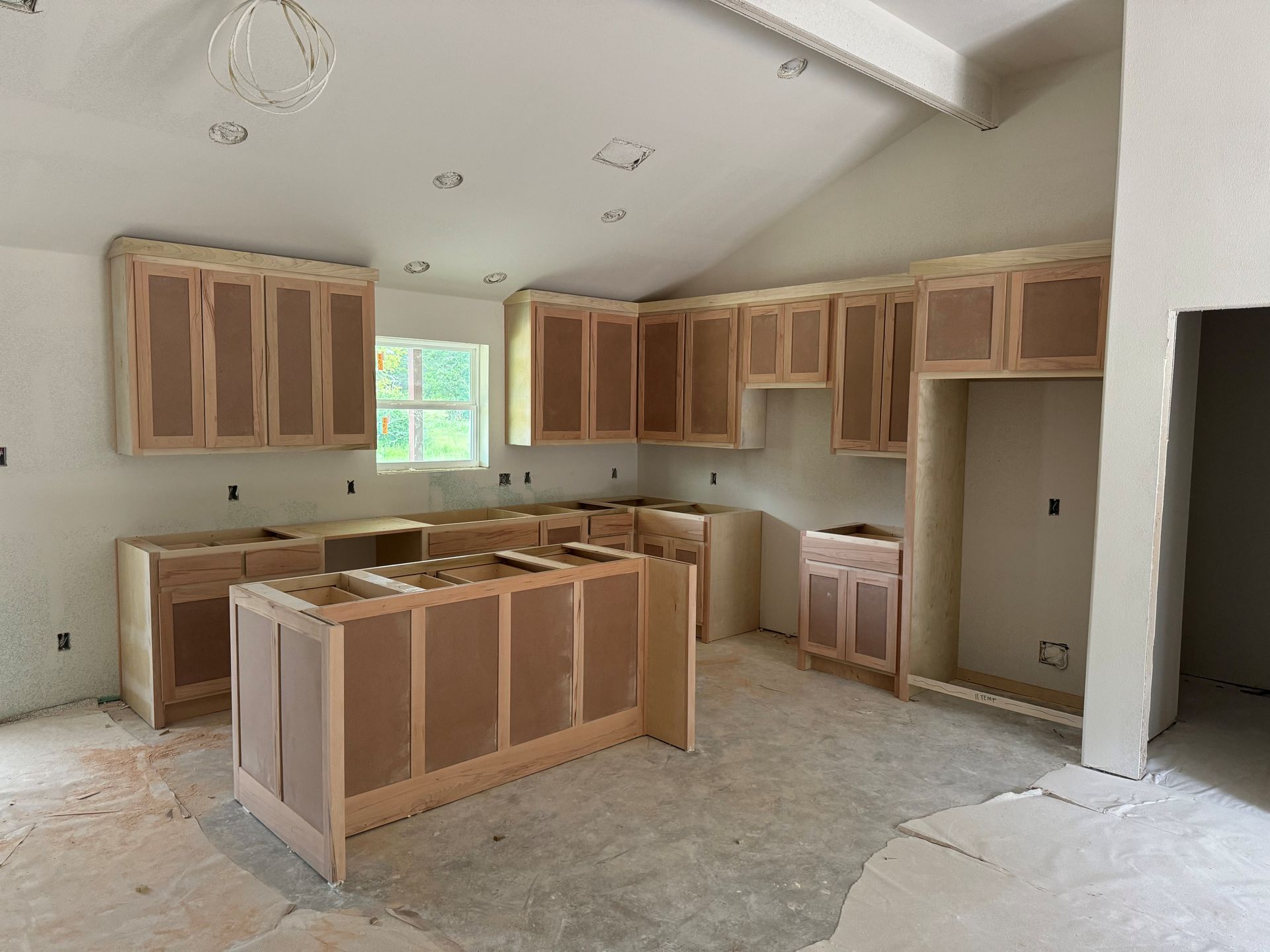 Unfinished kitchen with bare wood cabinets installed along the walls and a central island in a blank room.