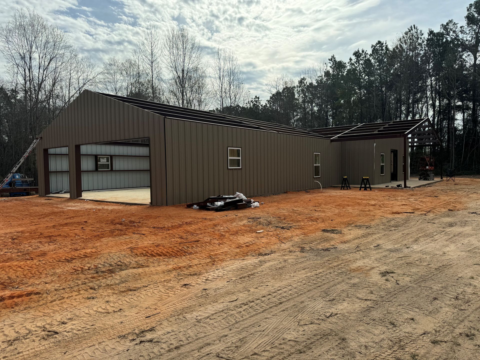 Large brown metal building under construction on a dirt lot, with trees and cloudy sky behind it