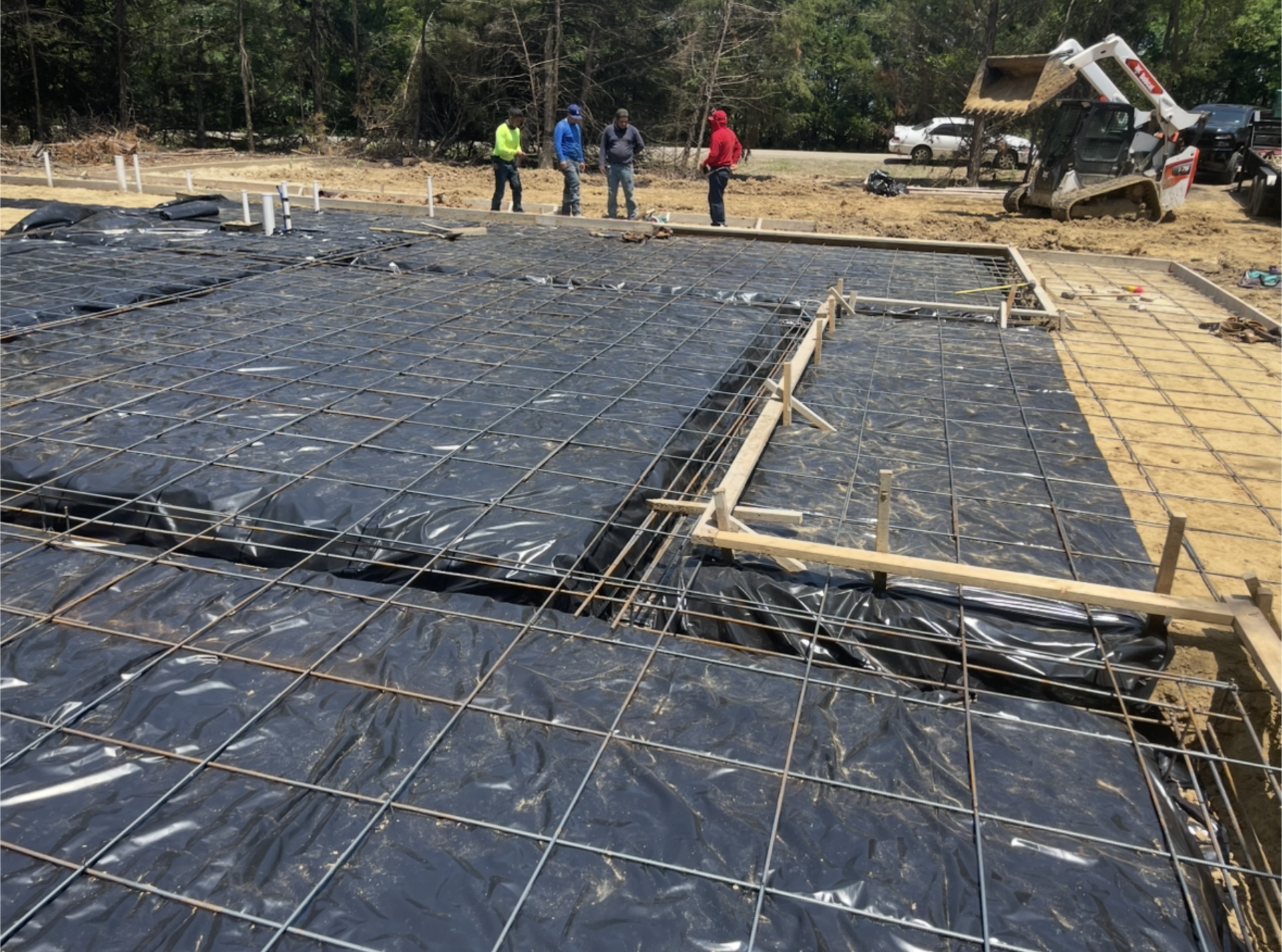 Construction site with rebar and black vapor barrier, workers in the background, and an excavator nearby